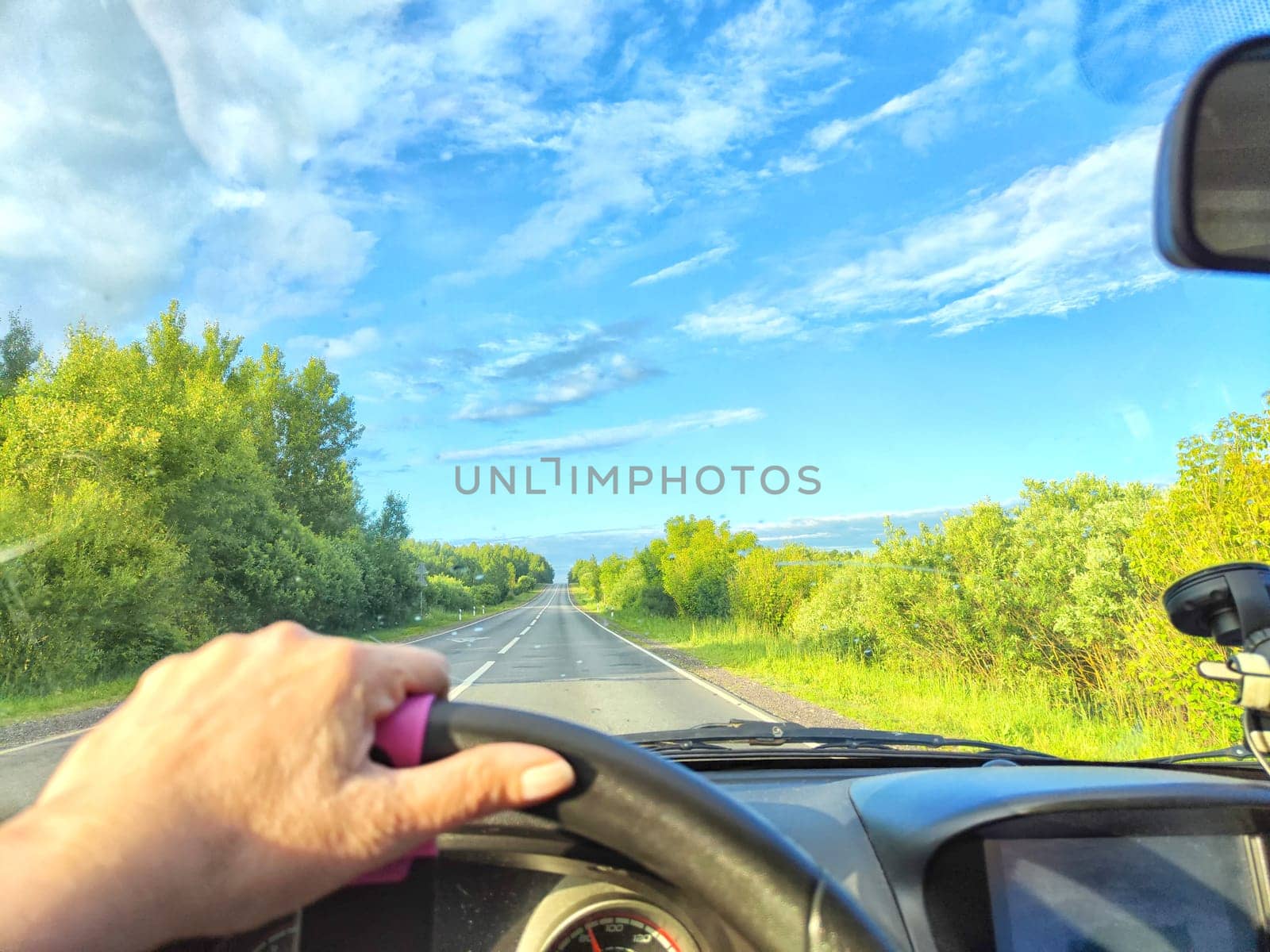 Car salon, windshield, hand of woman on steering wheel and landscape. View from seat of driver on nature with Road, trees, blue sky at a sunny day. Single trip of female traveler. Partial focus