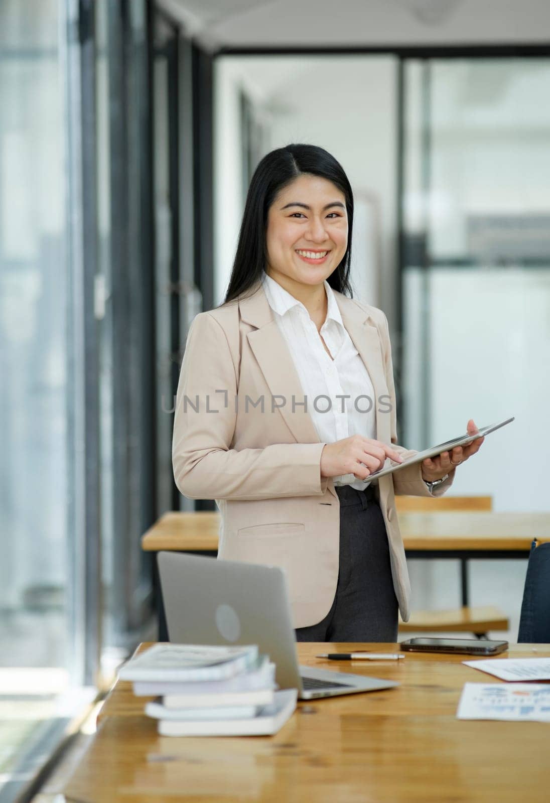 A professional and confident businesswoman smiling at the camera, standing by her desk with a laptop in a modern office.