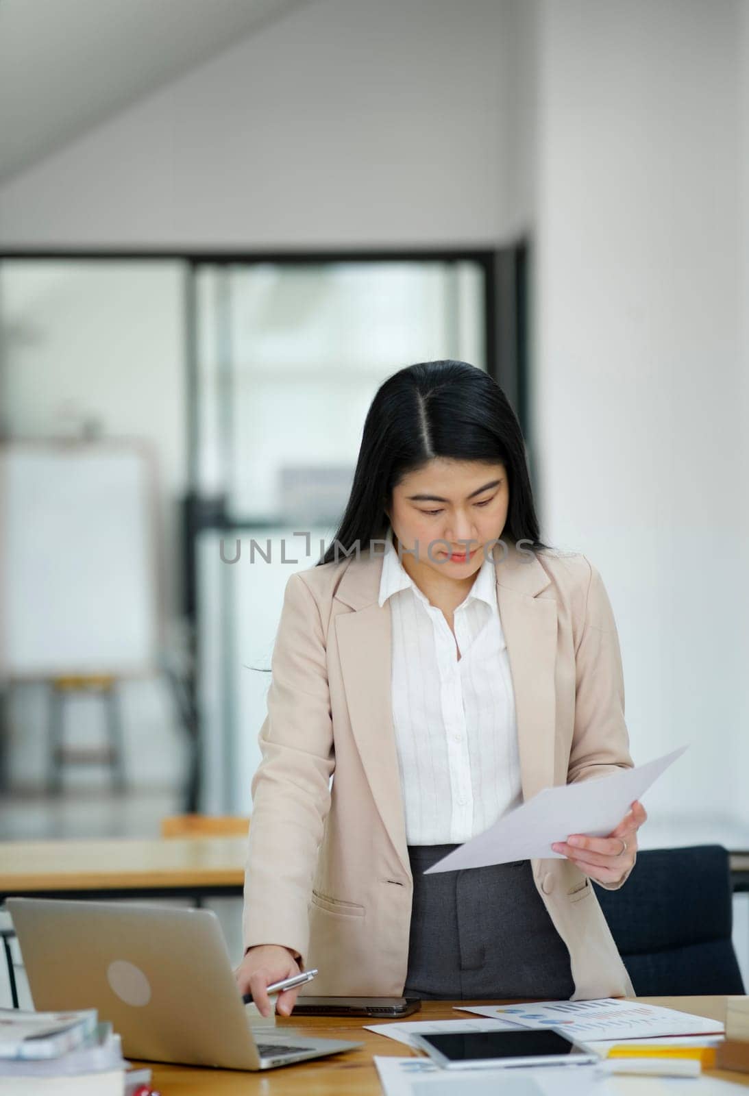 A focused businesswoman working on a laptop while holding a notebook in a bright office environment..