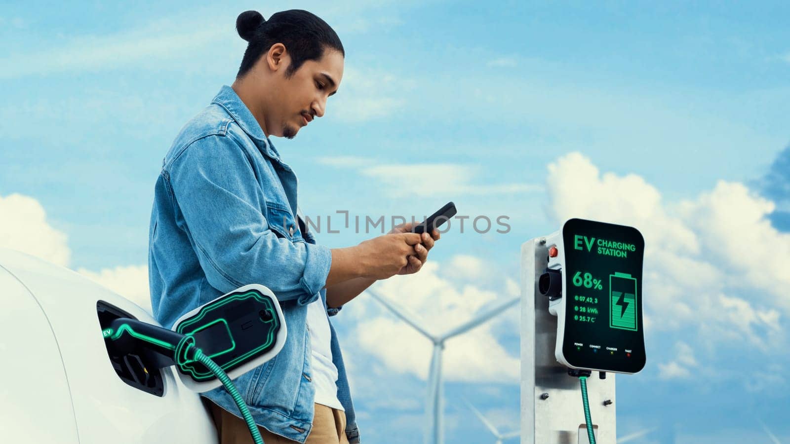 Asian man using smartphone while electric car recharging energy from EV charger from charging station at wind turbine farm. Alternative sustainable and clean energy utilization concept. Peruse