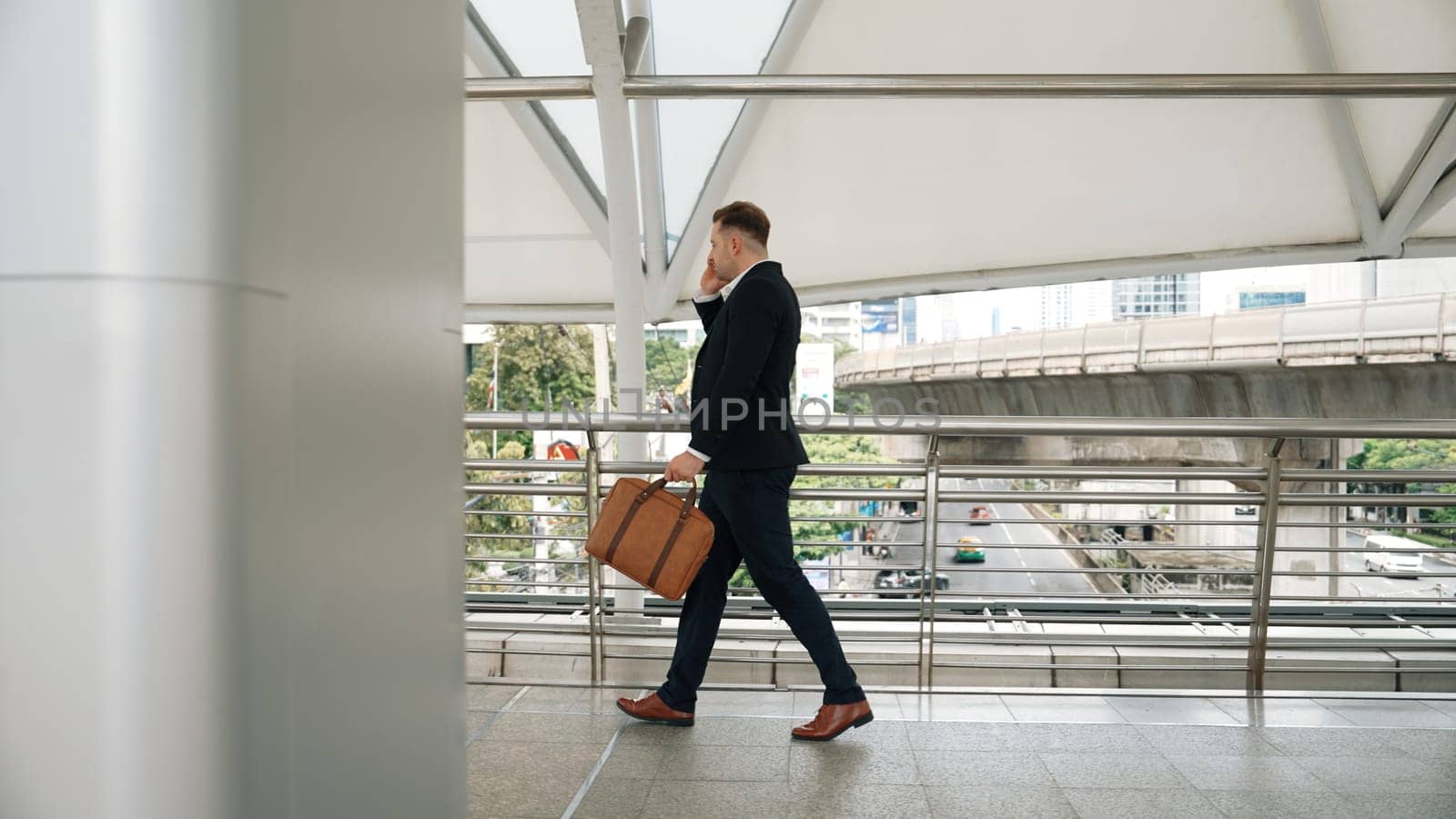 Smart caucasian businessman calling his colleague to plan financial strategy while walking to workplace. Side view of manager using mobile phone to communicate with marketing team. Lifestyle. Urbane.