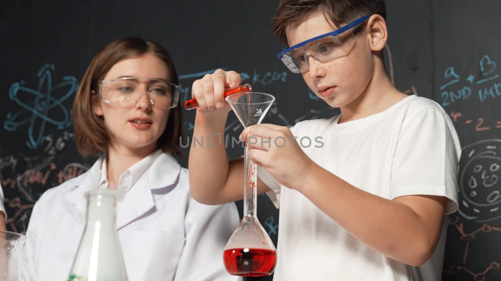 Close up of caucasian boy mixing chemical liquid while teacher giving advice. Instructor wearing lab suit looking for diverse student at table with beaker filled with colored solution. Erudition.