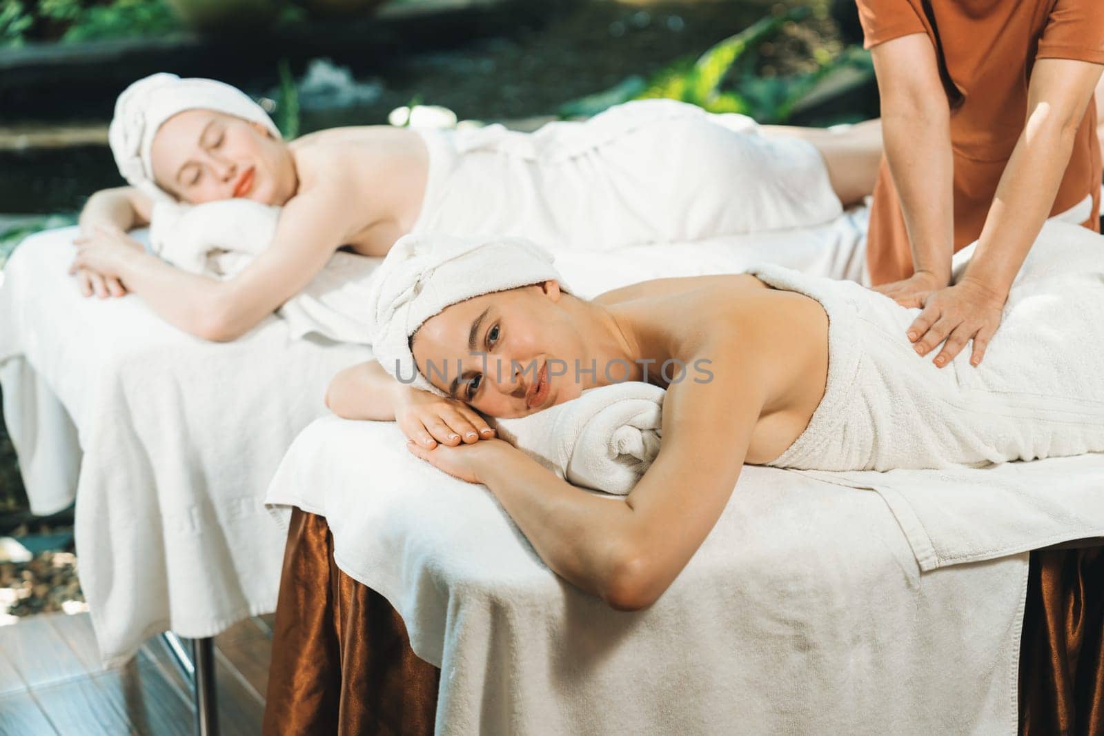 A portrait of two young attractive woman lie on bed during having back massage by a professional masseur at outdoor surrounded by peaceful natural environment. Healthy and beauty concept. Tranquility.
