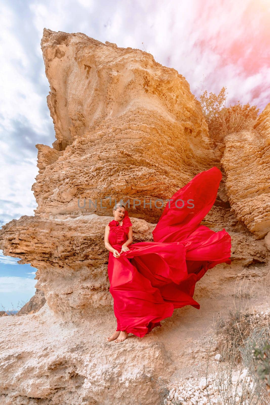 woman red silk dress stands by the ocean, with mountains in the background, as her dress sways in the breeze