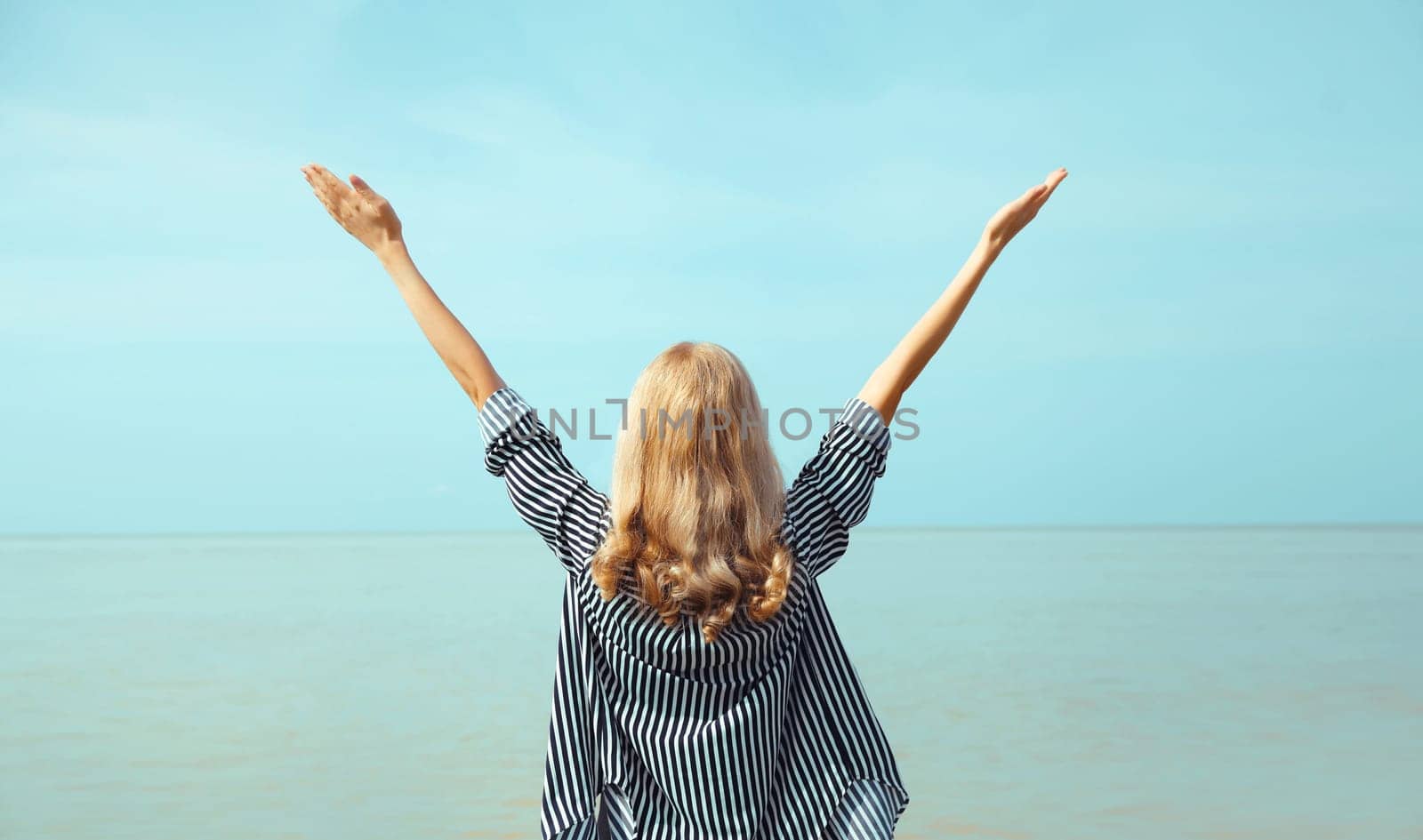Summer vacation, happy young woman raising her hands up on the beach on sea coast and blue sky background