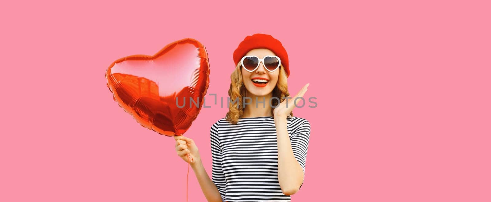 Cute portrait of happy cheerful smiling young woman with red heart shaped balloon wearing french beret hat on pink studio background