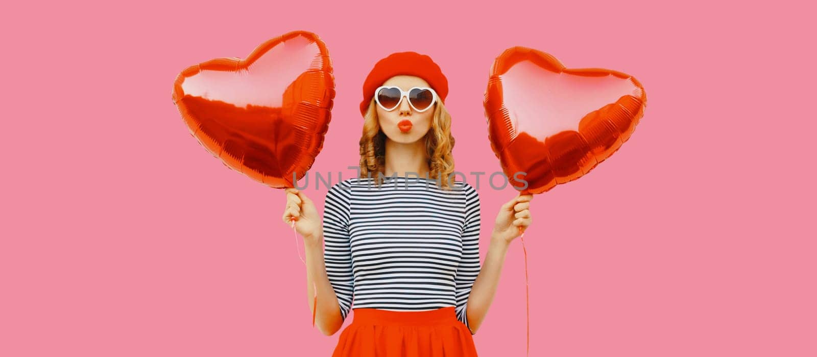 Sweet portrait of beautiful happy young woman with red heart shaped balloon blows kiss in french beret hat on pink studio background