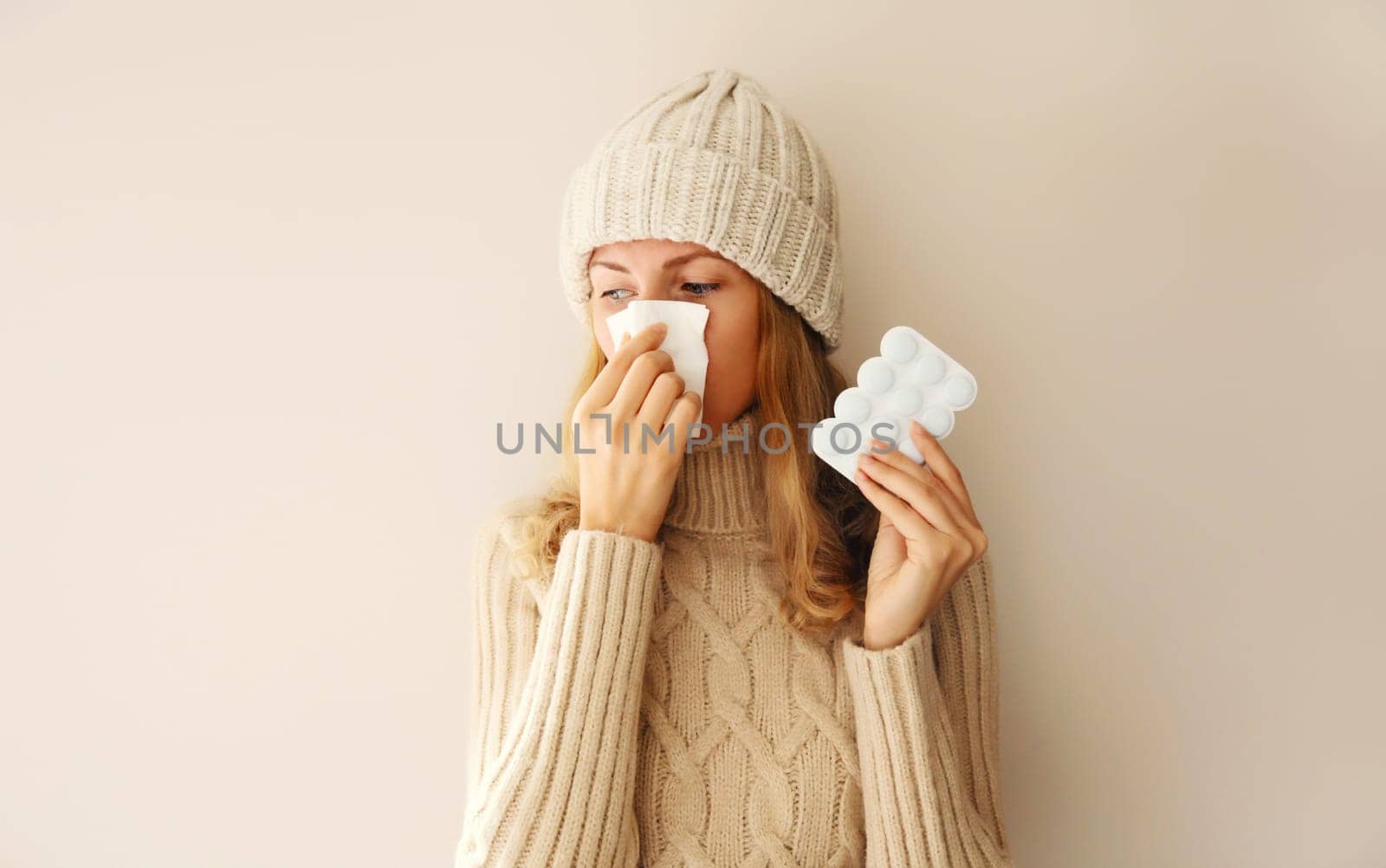 Sick woman sneezing blow nose using tissue and holding pills wearing warm soft knitted clothes, hat and sweater on beige studio background