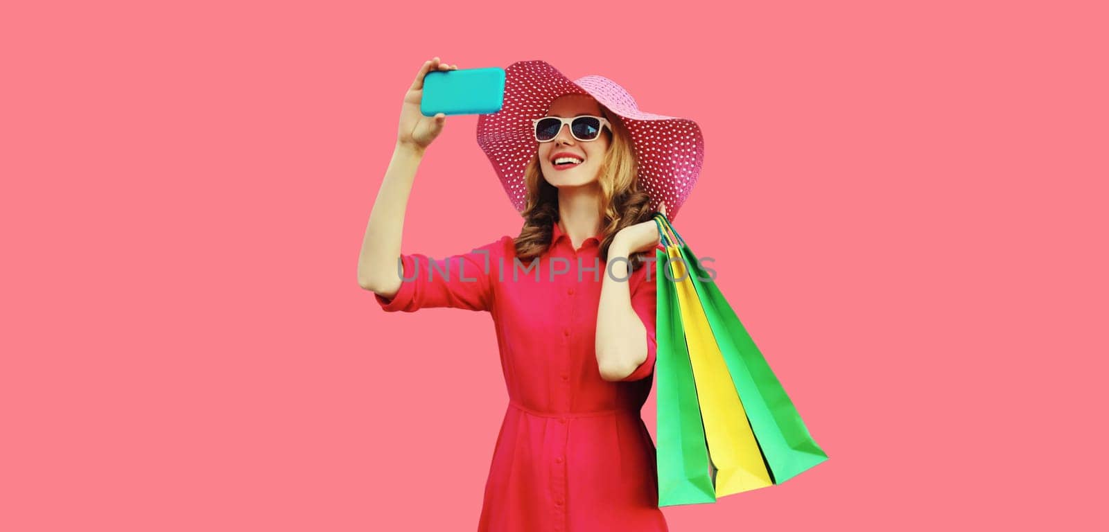 Beautiful happy smiling young woman model taking selfie with mobile phone holding colorful shopping bags in summer straw hat, dress on pink background