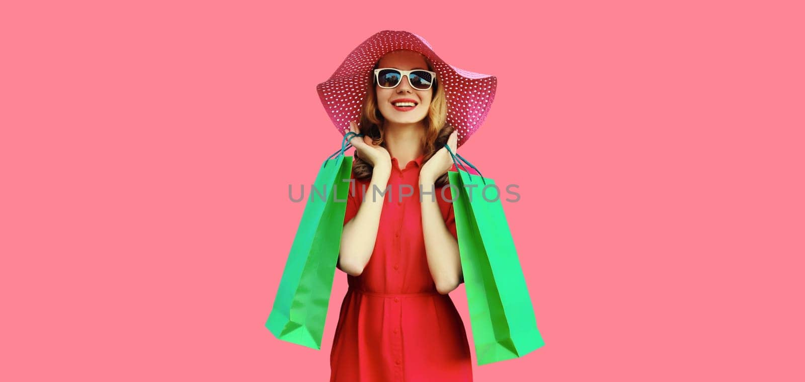 Portrait of beautiful happy smiling young woman model posing with colorful shopping bags in summer straw hat, dress on pink studio background