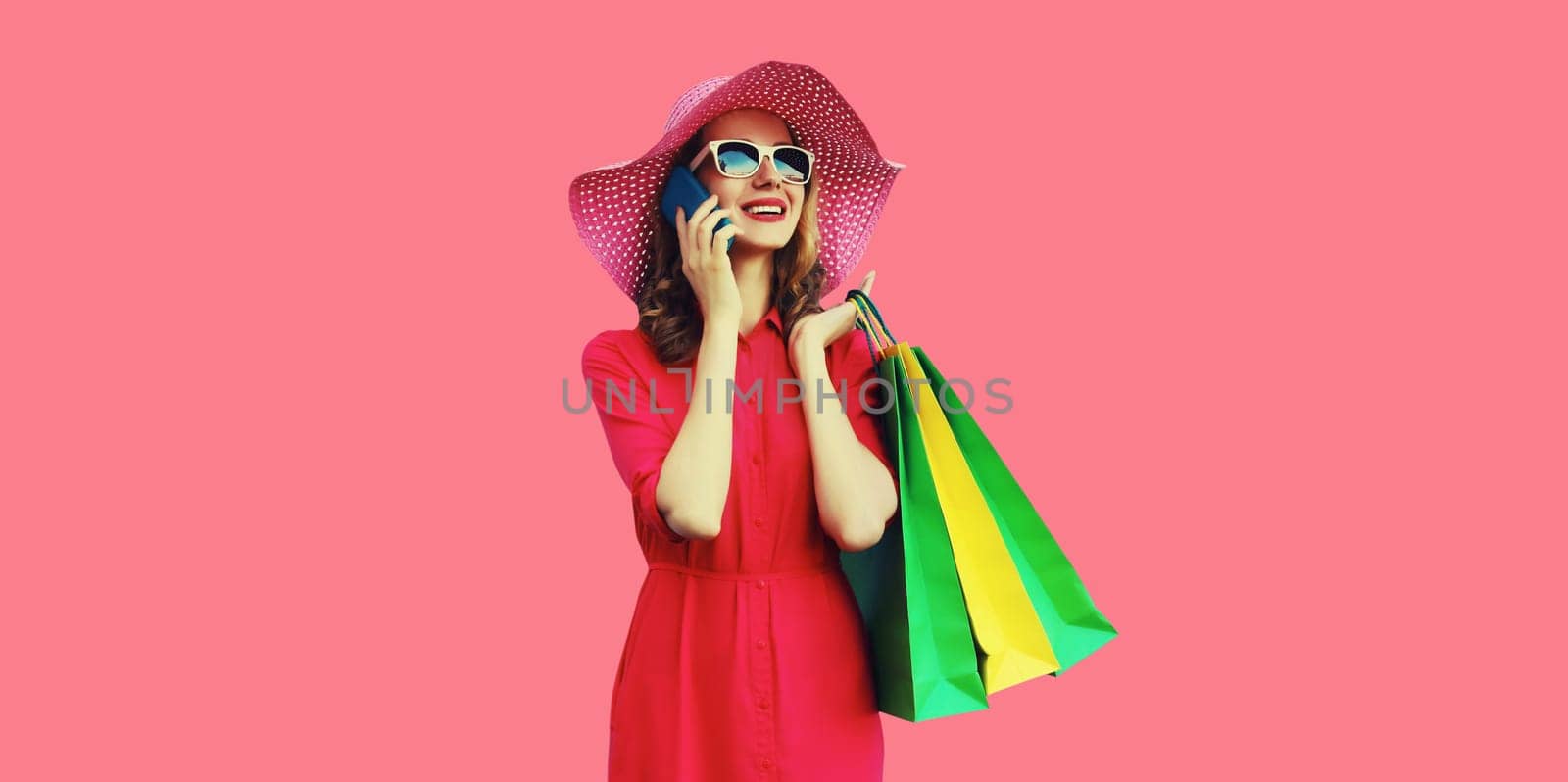 Beautiful happy young woman calling on mobile phone with shopping bag looking away in summer hat on pink studio background