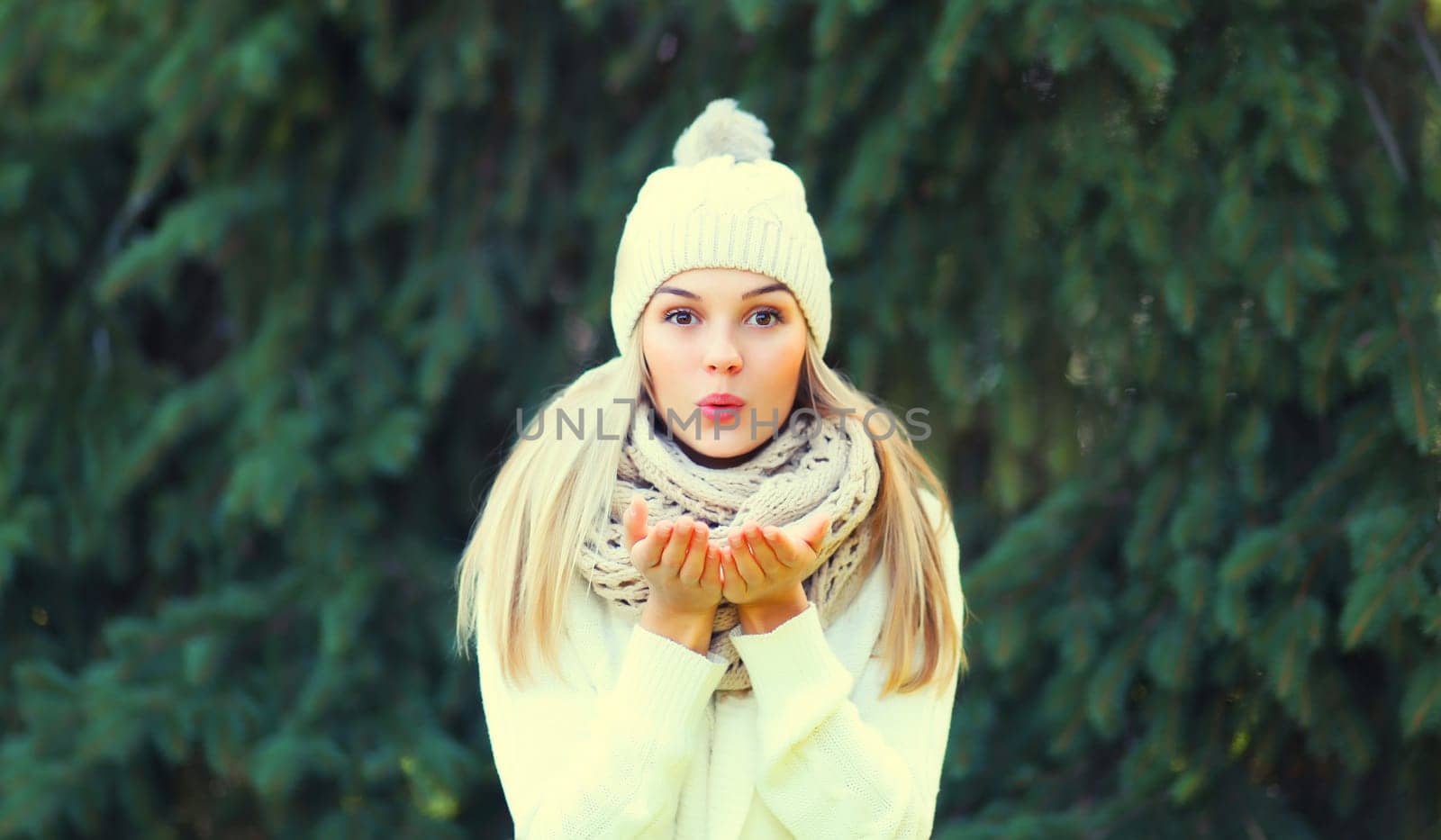 Winter portrait of happy cheerful blonde young woman blowing snowflakes from hands in hat, sweater in forest on christmas tree background