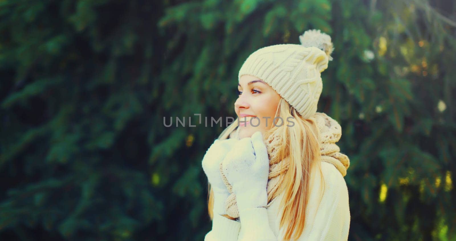 Winter portrait of happy smiling young woman wearing hat, knitted sweater and scarf in forest on christmas tree background