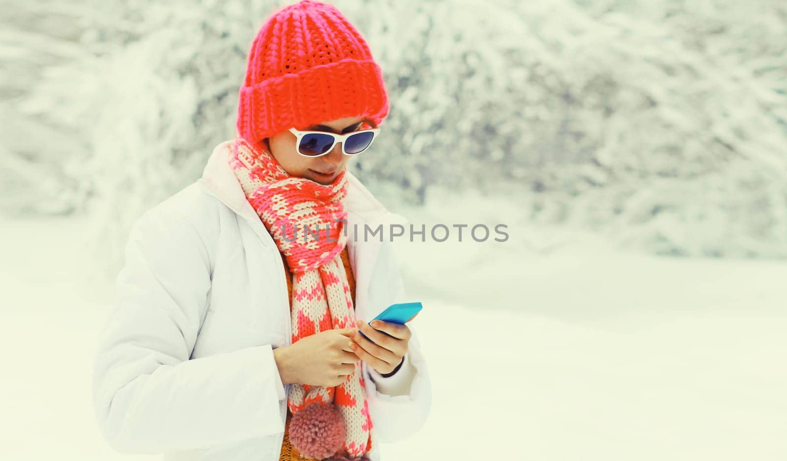 Winter portrait of happy young woman with mobile phone in knitted hat, scarf and jacket in snowy park