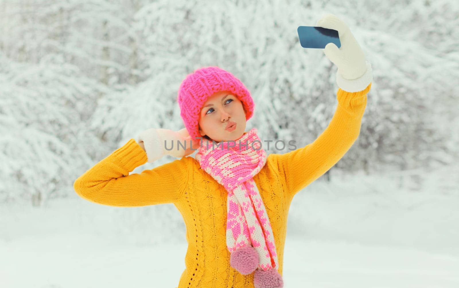 Winter portrait of happy cheerful young woman taking selfie with phone having fun in knitted sweater, hat and scarf in snowy park