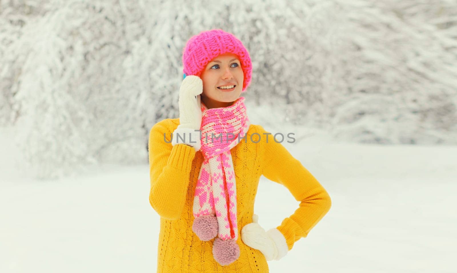 Happy young woman calling on smartphone looking away in colorful knitted hat, scarf and sweater in snowy park