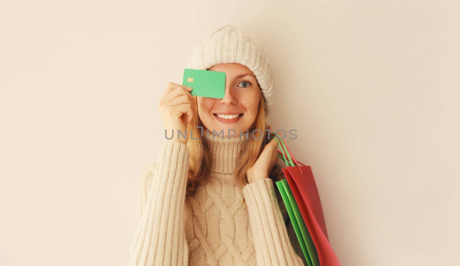 Portrait of happy smiling young woman holding plastic credit bank card with shopping bags in winter knitted hat and sweater on beige studio background