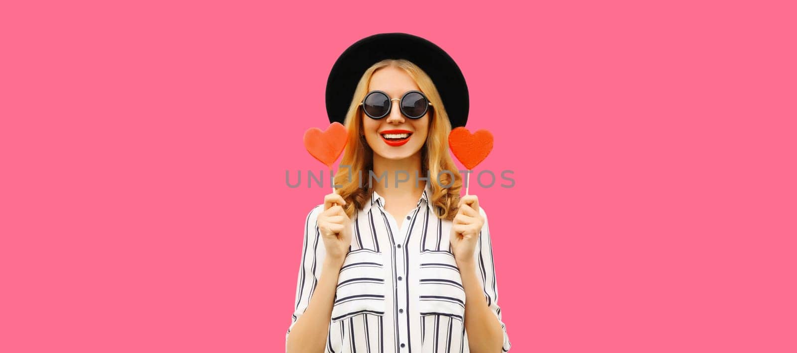 Portrait of happy cheerful young woman with sweet red heart shaped lollipop on stick in black round hat on pink studio background