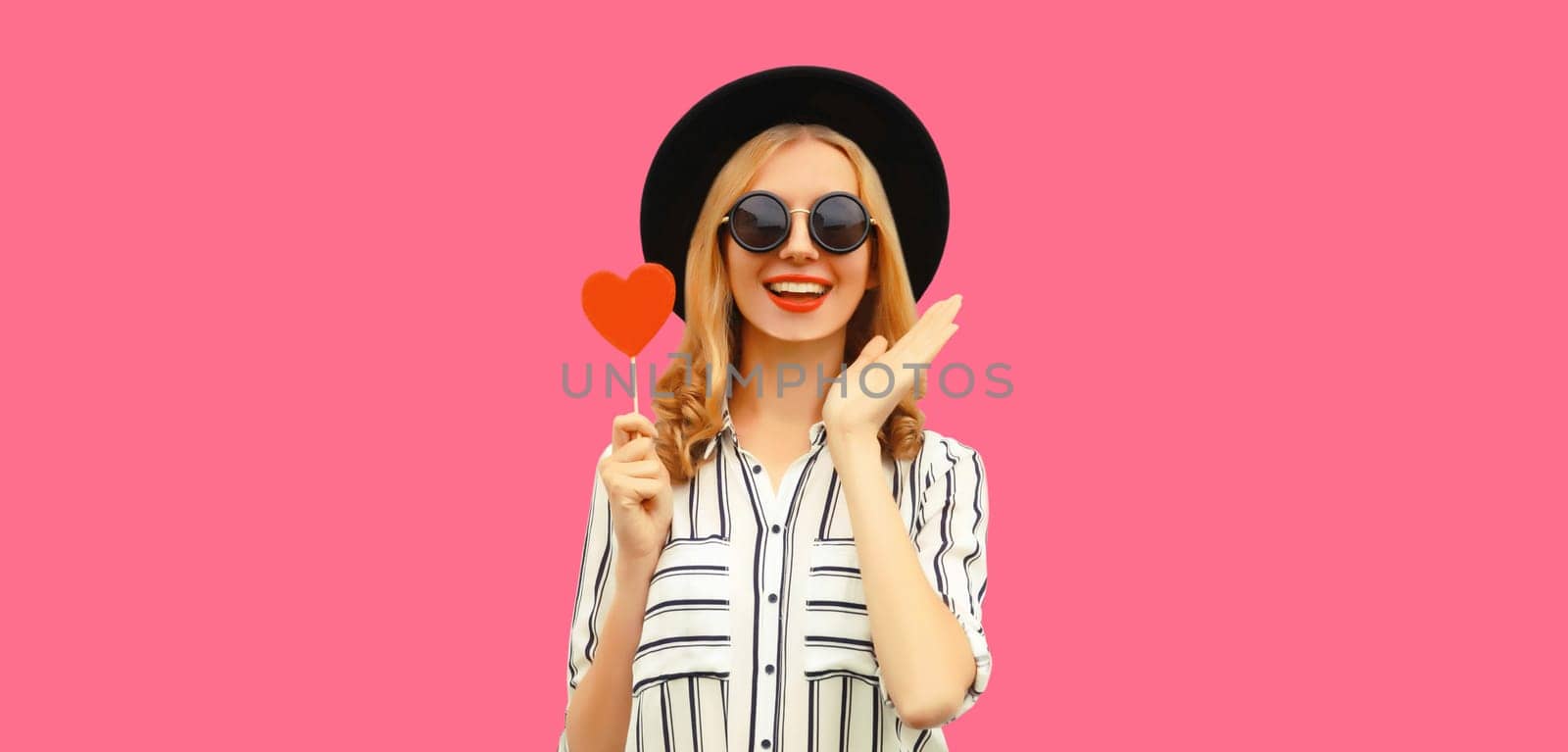 Portrait of happy cheerful young woman with sweet red heart shaped lollipop on stick in black round hat on pink studio background