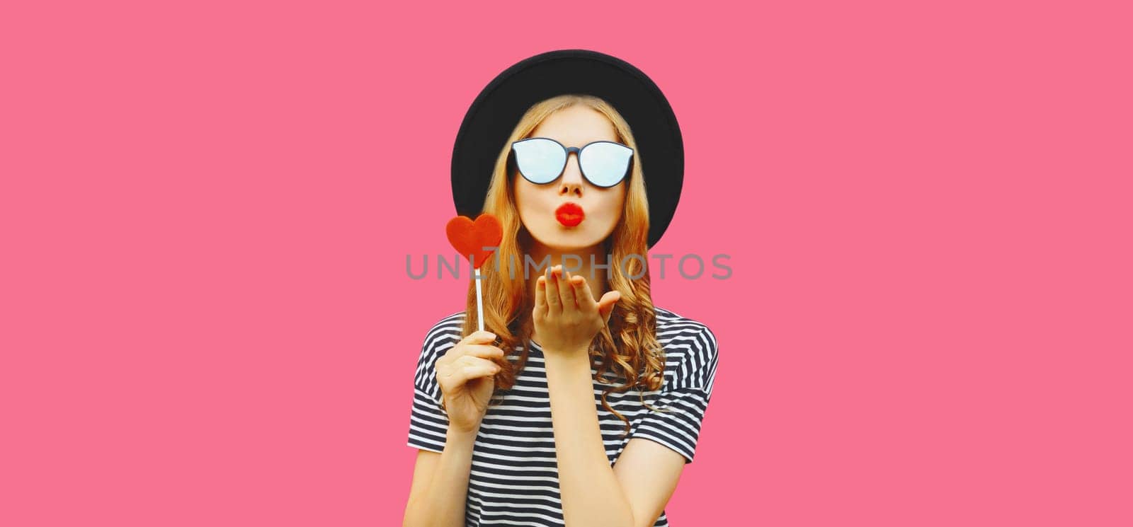 Portrait of happy beautiful young woman blowing her lips sends kiss with sweet red heart shaped lollipop on stick in black round hat on pink studio background