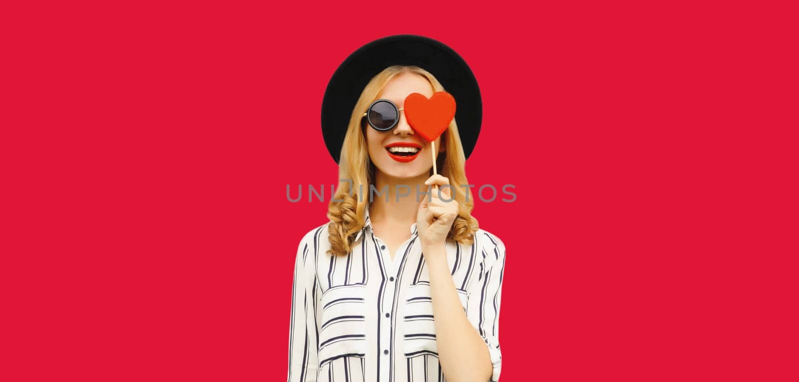 Portrait of happy cheerful young woman with sweet red heart shaped lollipop on stick in black round hat on pink studio background