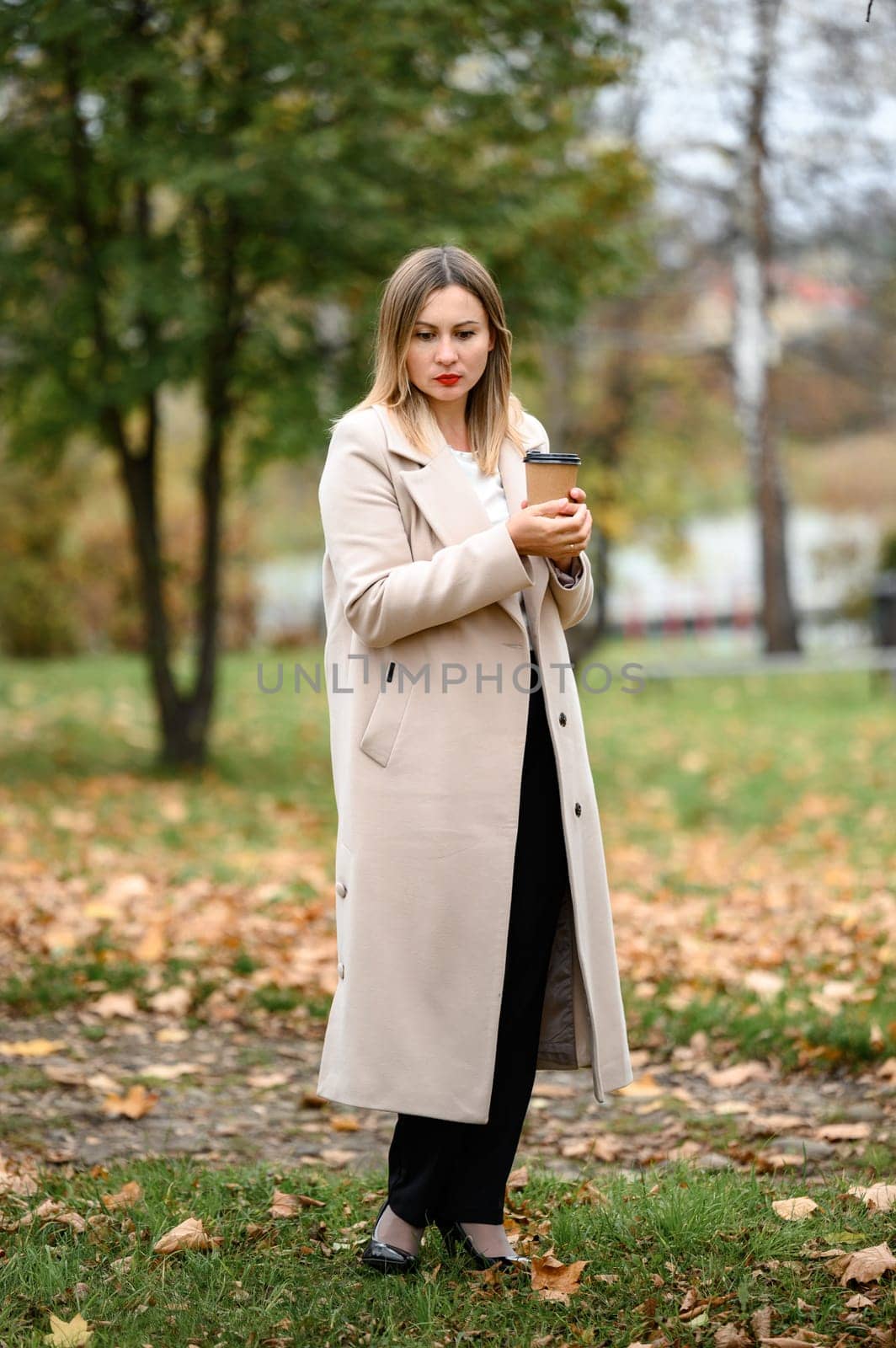 A young girl in a long autumn coat drinks coffee in an autumn park. by Niko_Cingaryuk