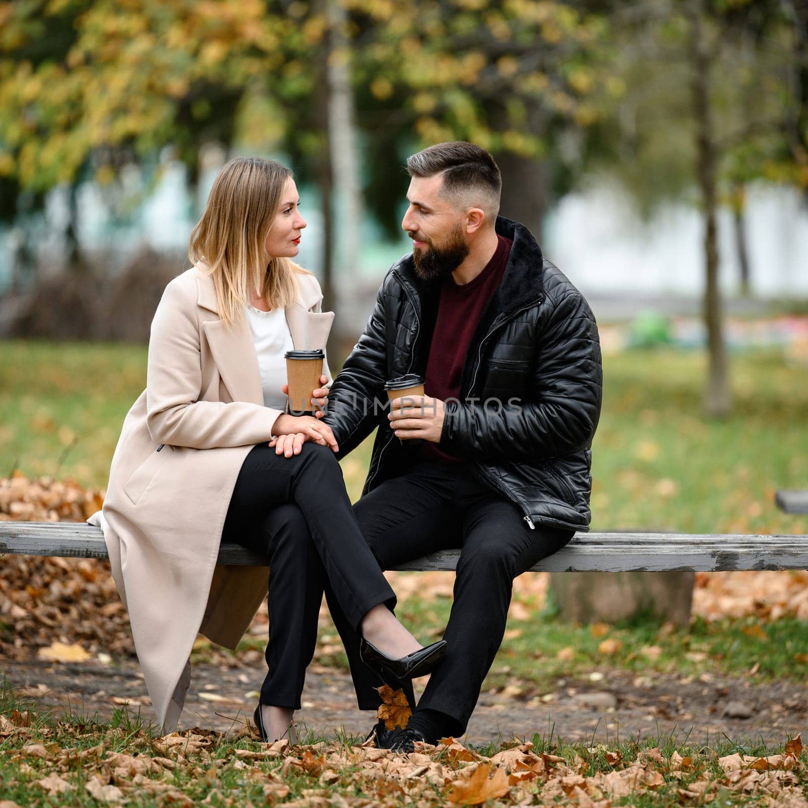 A couple, a man and a woman, are sitting in the park on a bench with coffee in their hands. by Niko_Cingaryuk