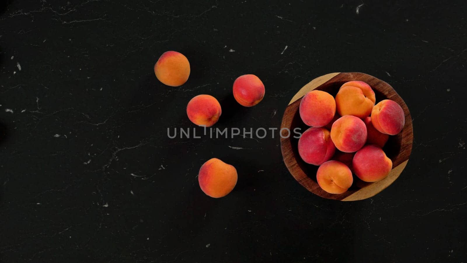 Ripe apricots in small wooden bowl, some scattered on black marble like desk next to it. View from above, space for text left
