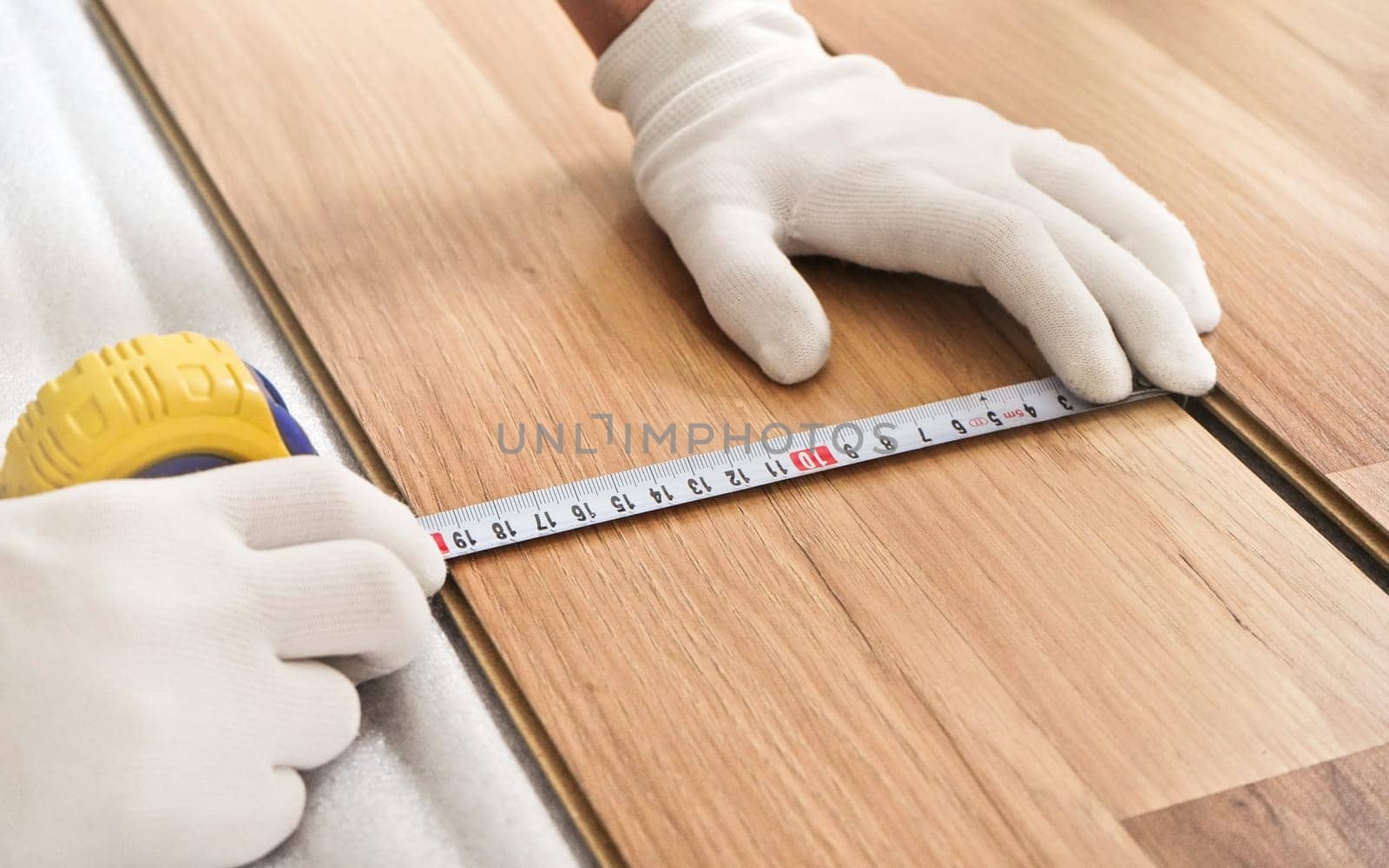 Installing laminated floor, detail on man hands in white gloves holding measuring tape over wooden tile
