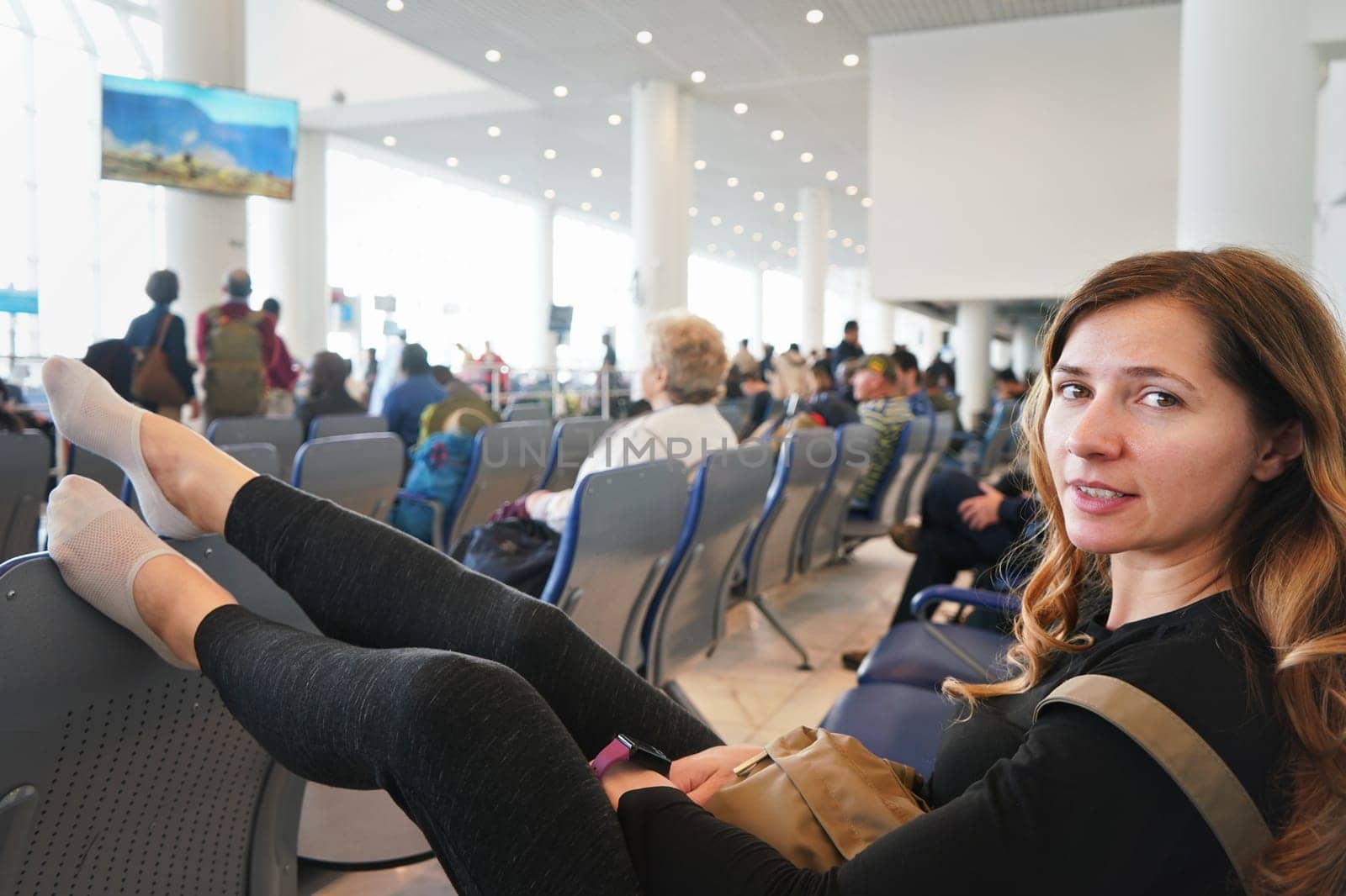 Young woman sitting at airport hall, looking tired after waiting couple of hours for connecting flight in early morning, legs on seat in front of of her, more blurred passengers in background