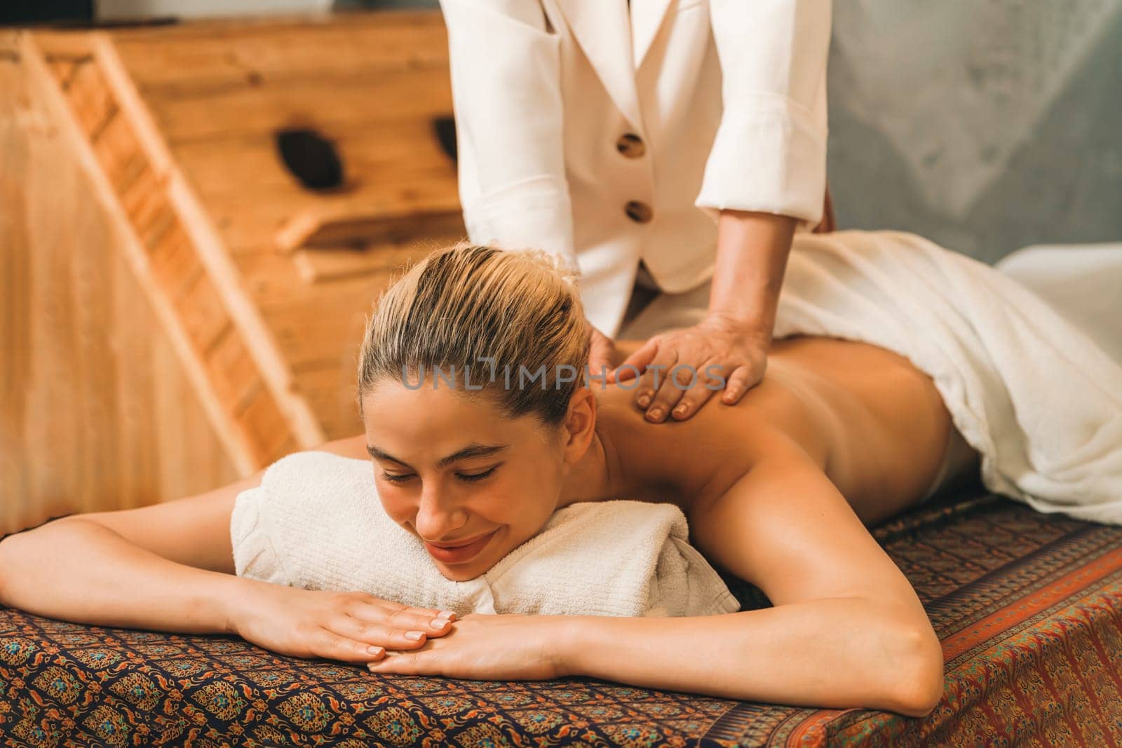 Beautiful young woman lies on spa bed in front of wooden sauna cabinet during having back massage. Attractive women in white towel in warm sauna room. Relaxing and healthy concept Tranquility.