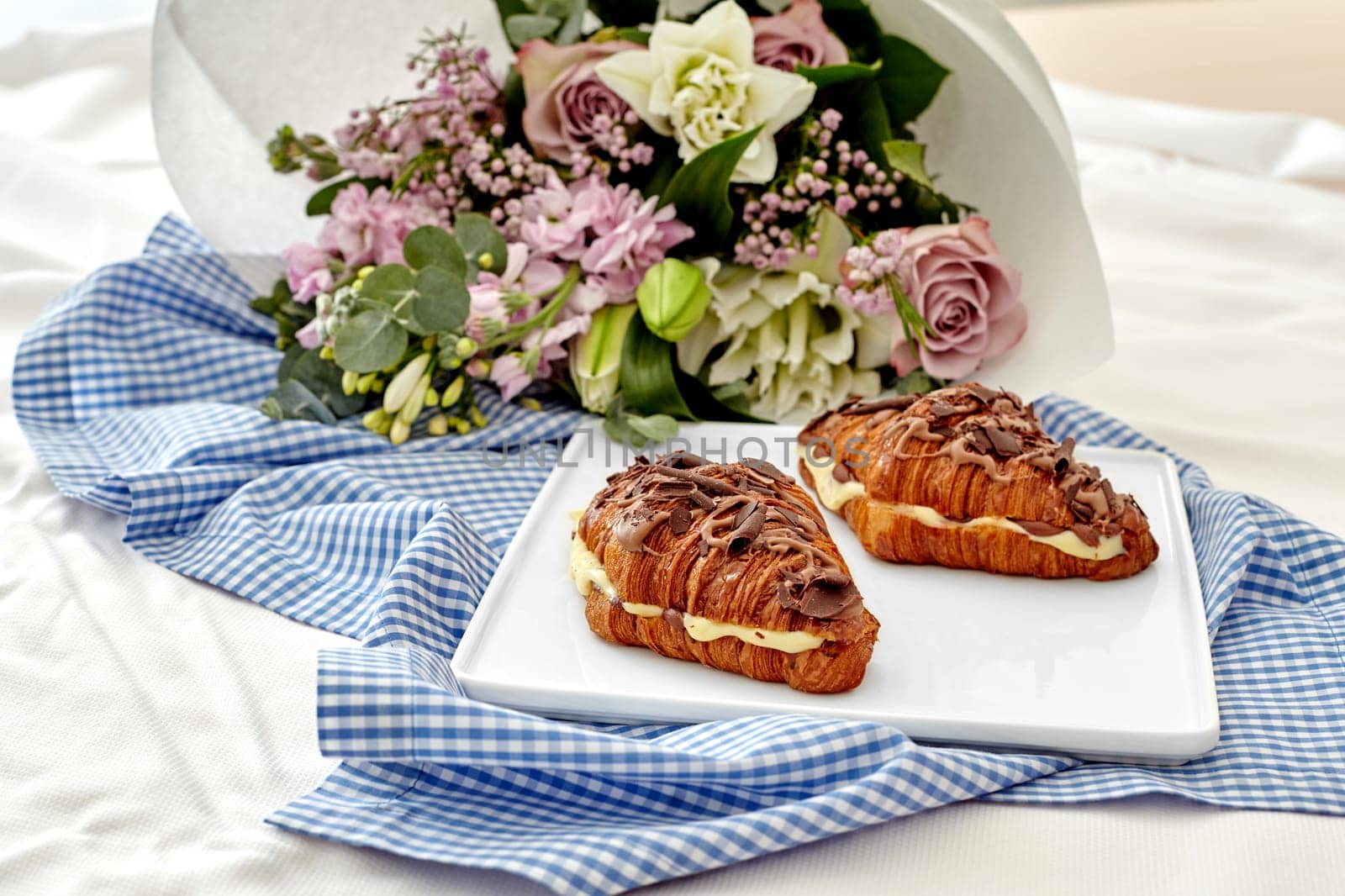 Romantic setting featuring chocolate drizzled puff French croissants sandwiches stuffed with delicate custard and fresh flower bouquet on white blue gingham cloth