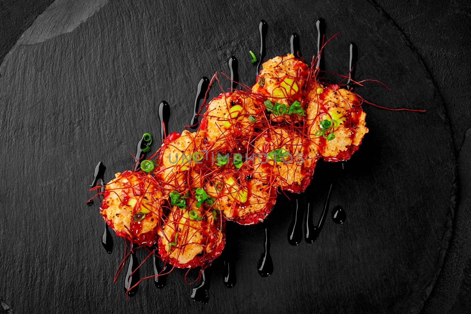 Top view of appetizing red rice sushi rolls topped with seared scallop cheese caps drizzled with spicy unagi sauce garnished with togarashi threads and green onions presented on black slate board