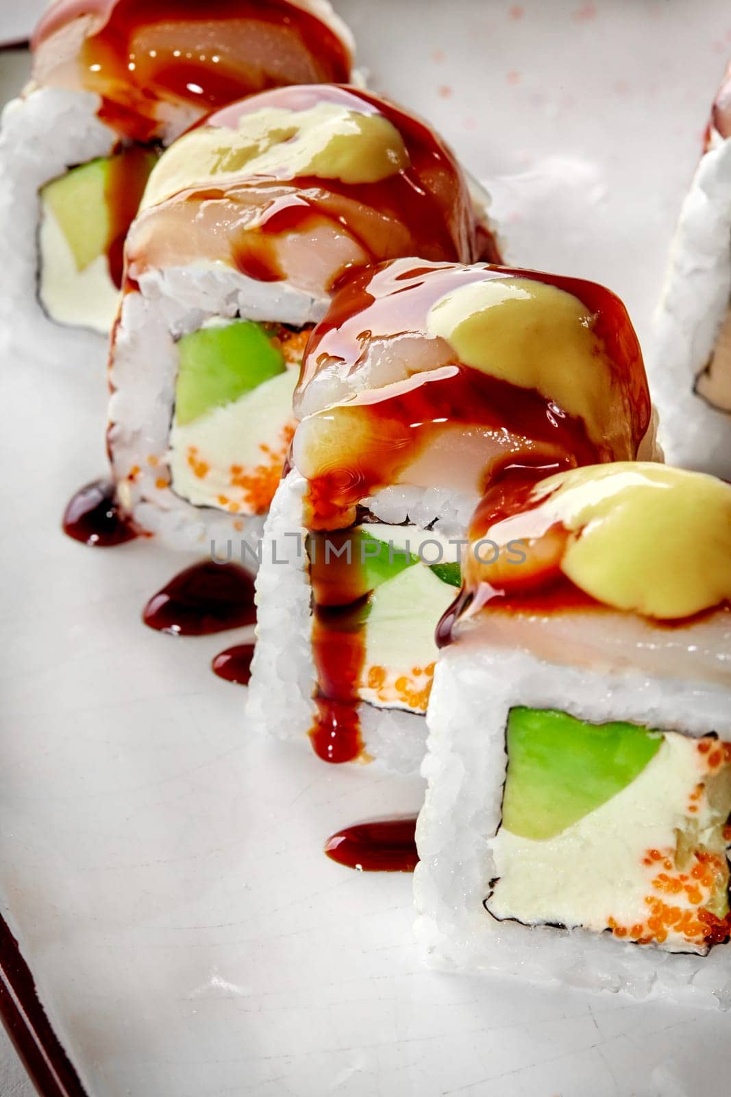 Closeup of sushi rolls filled with cream cheese, tobiko and avocado topped with sea scallop and drizzled with spicy mayo and savory unagi sauce, served on plate. Japanese cuisine