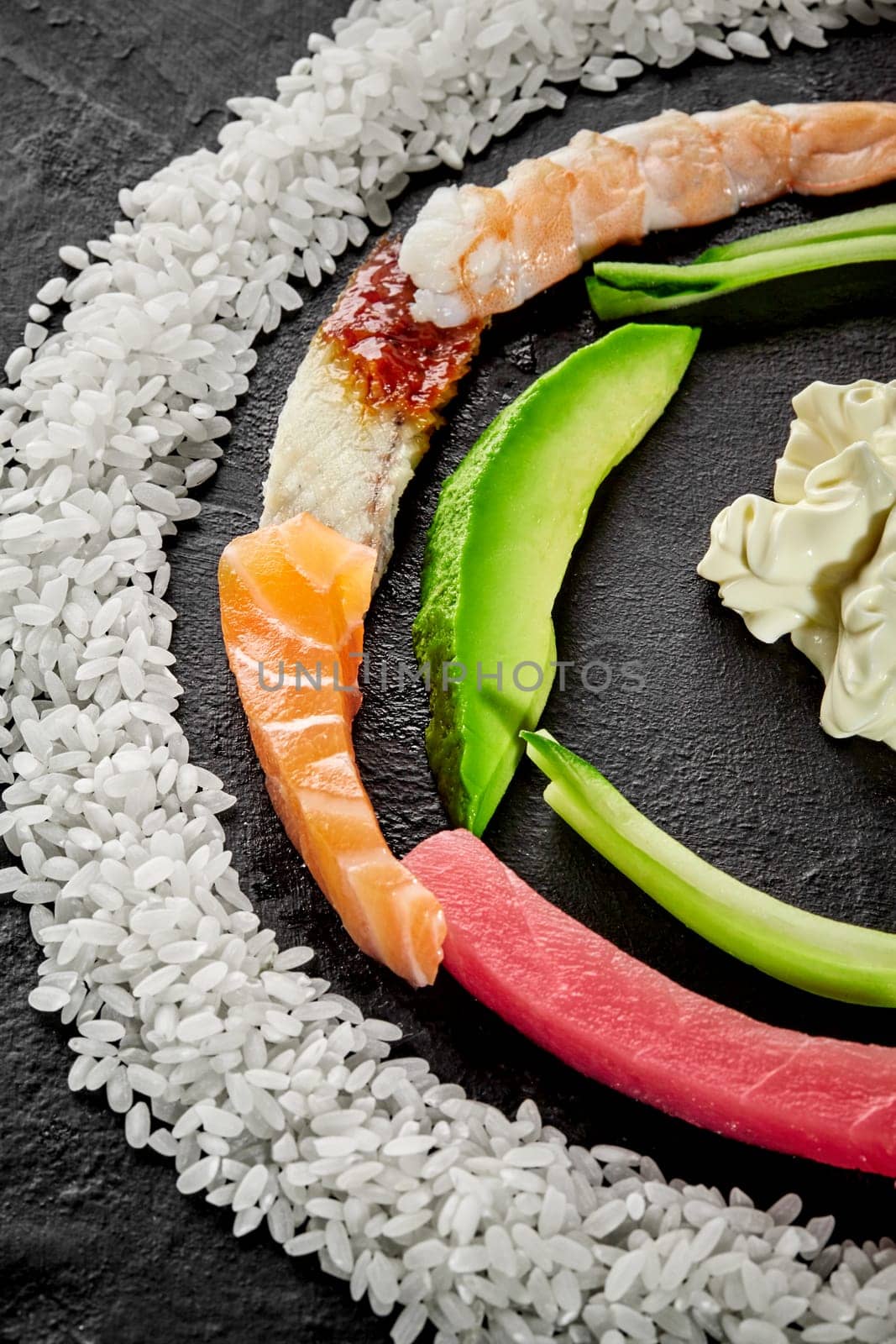 Closeup of colorful half circle of slices of fresh raw salmon, tuna, eel, shrimps, ripe avocado, cucumber, white rice and Japanese mayonnaise on black textured background. Ingredients for making sushi