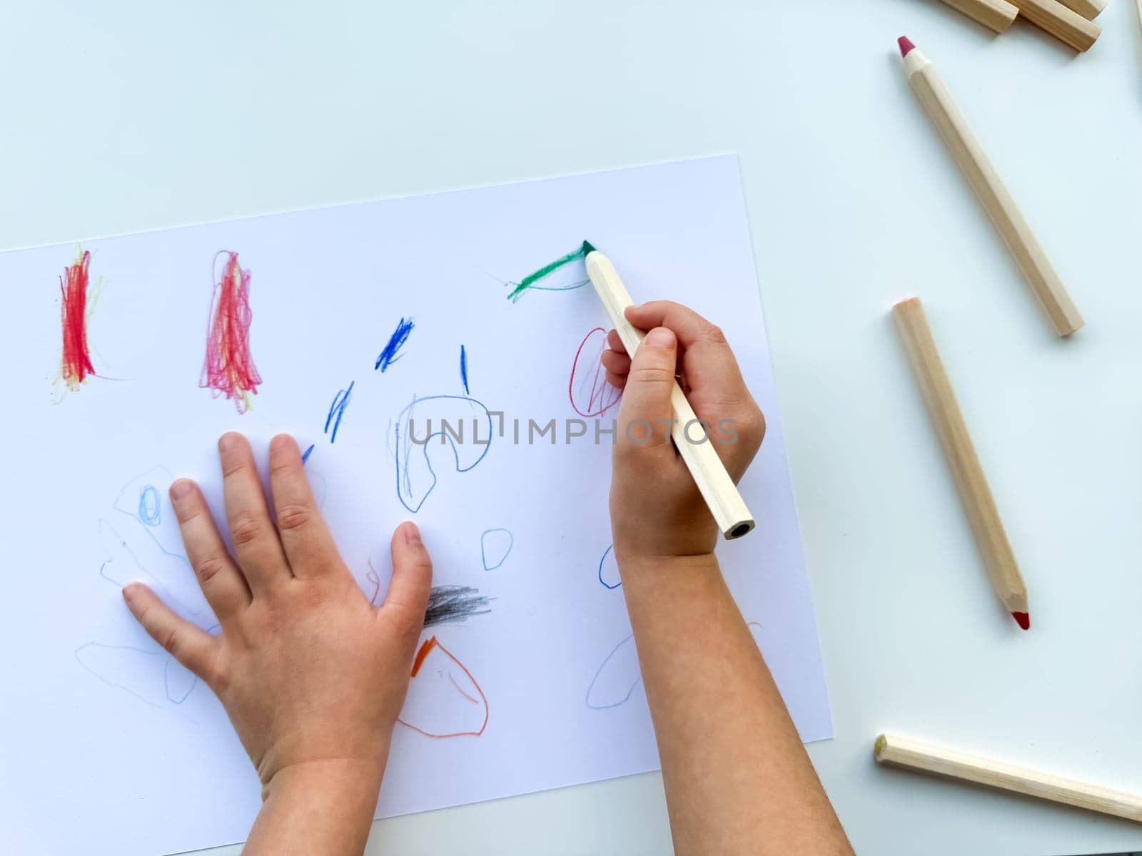 small child draws with colored pencils on paper on white table. High quality photo