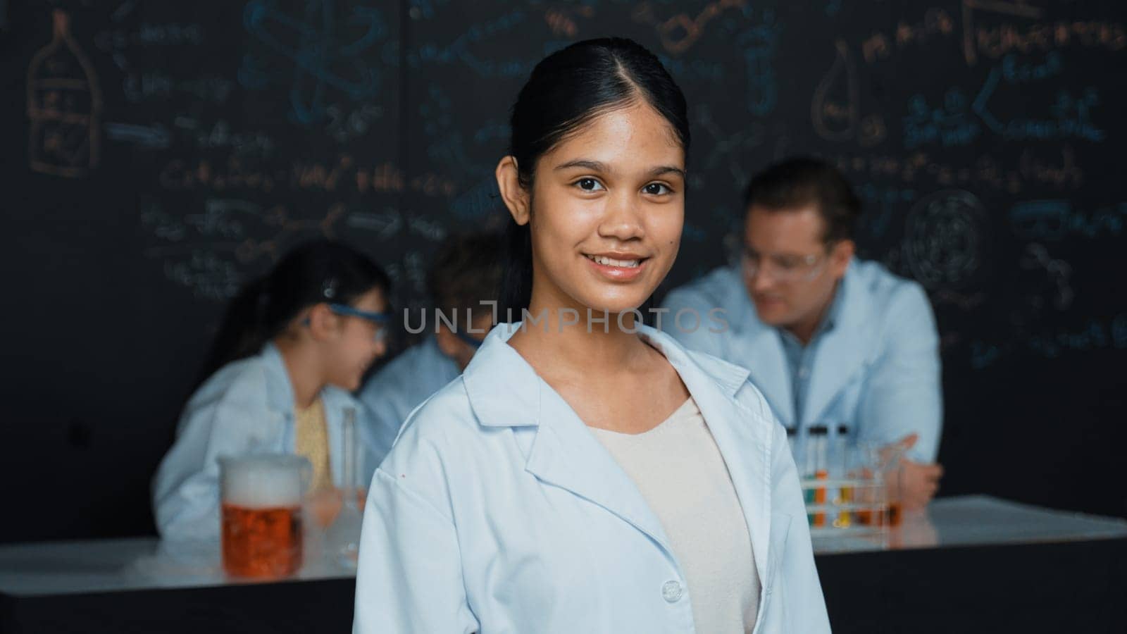 Girl looking at camera with arm folded while people doing experiment at laboratory. Cute student standing blackboard with chemical theory with blurring background at STEM science class. Edification.