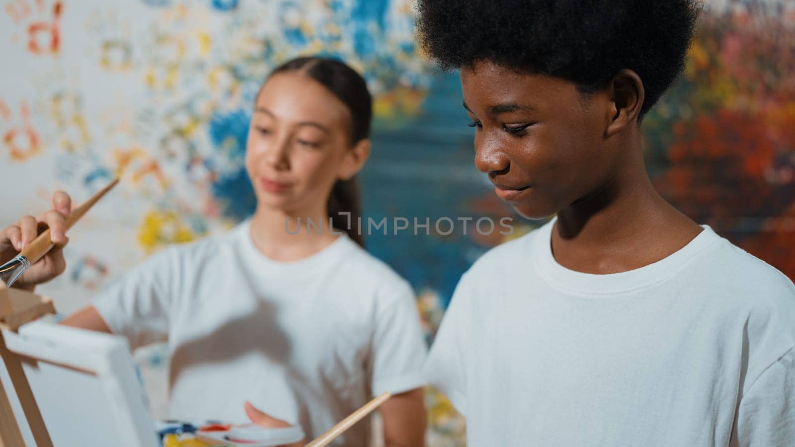 Diverse happy children paint canvas together with colorful stained wall. Smart multicultural student drawing together in art lesson while wearing white shirt. Creative activity concept. Edification.