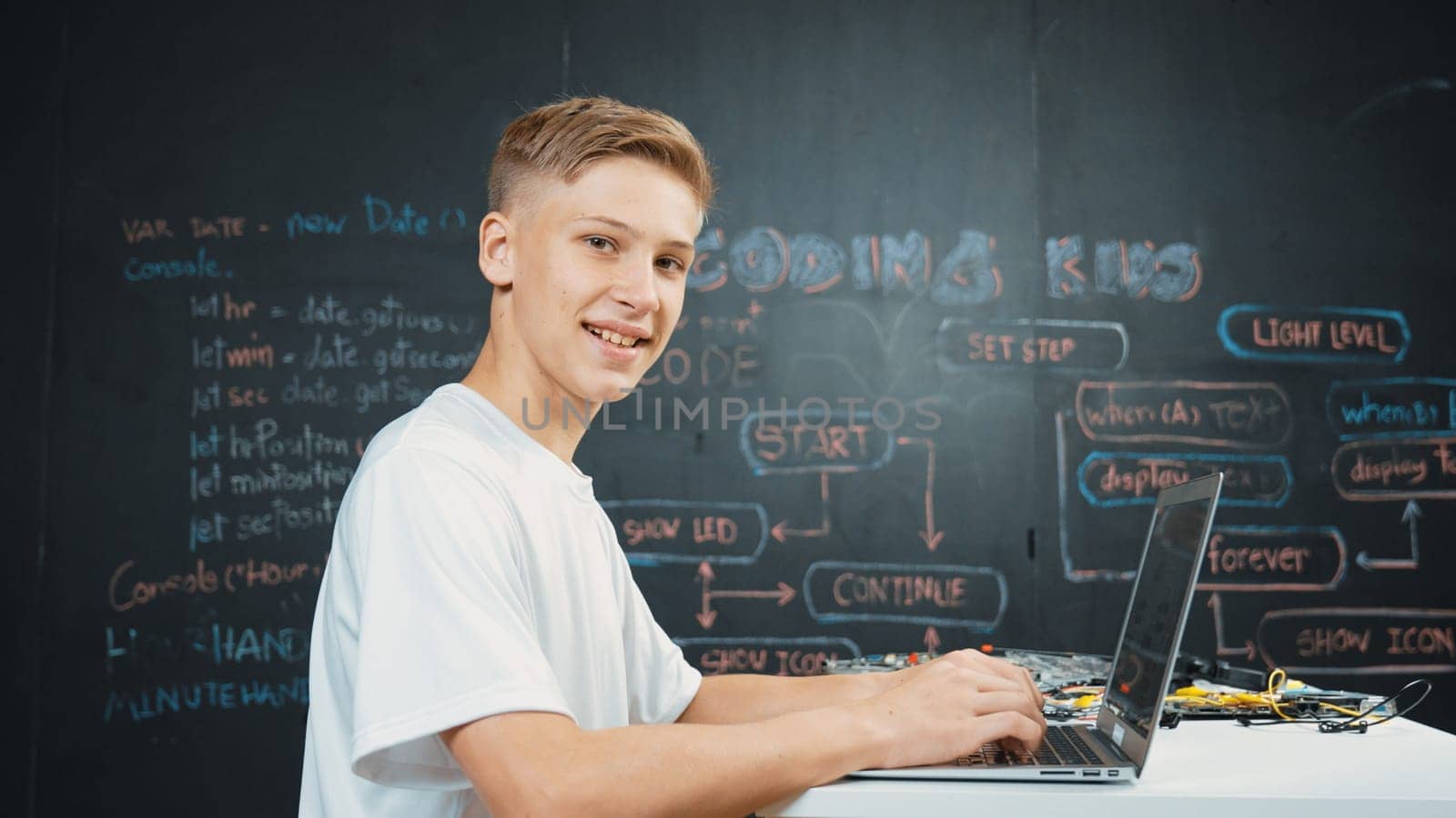 Caucasian boy coding and programing system while looking at laptop screen and camera with confident. Attractive academic student using computer analysis data while sitting at blackboard. Edification.
