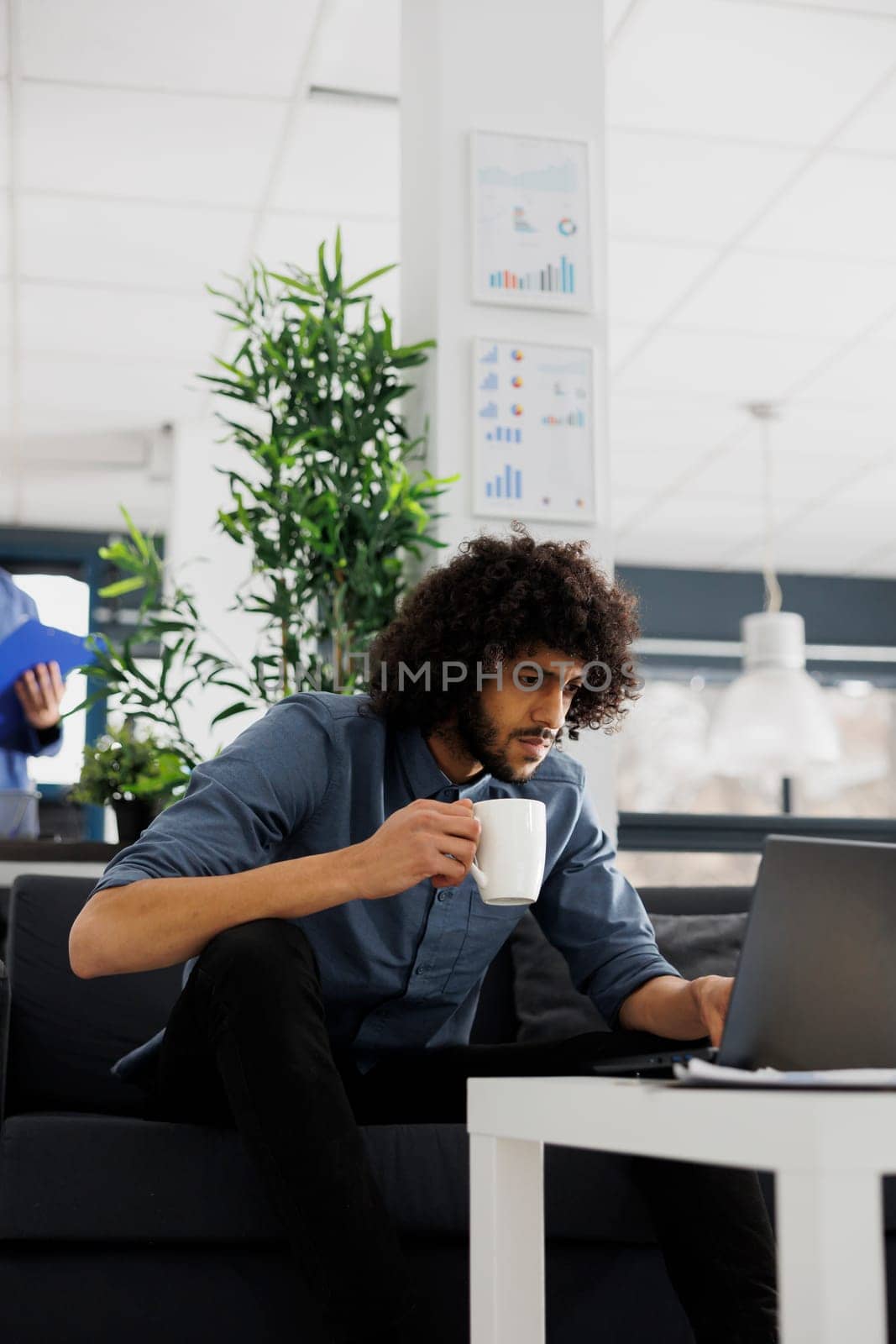 Arab company employee drinking coffee and reading customer email on laptop in office. Business start up focused entrepreneur holding tea mug and managing project in coworking space