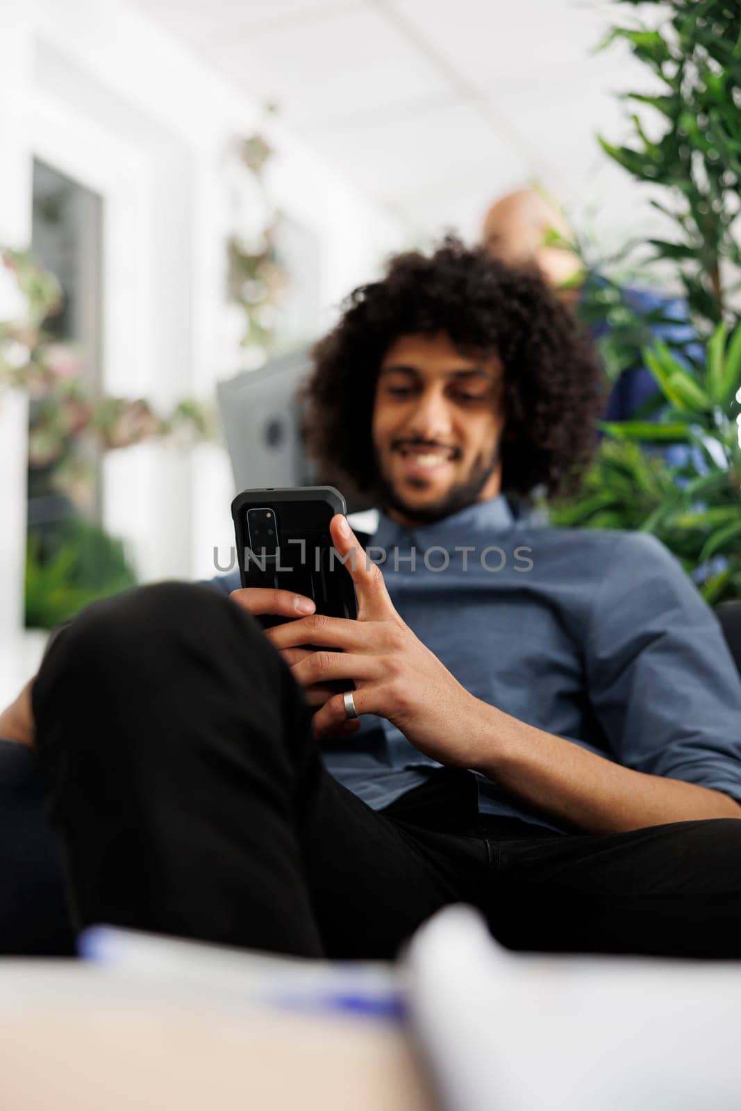 Smiling businessman relaxing in office, using smartphone to send message. Young arab executive manager texting in social media on mobile phone while having break in coworking space