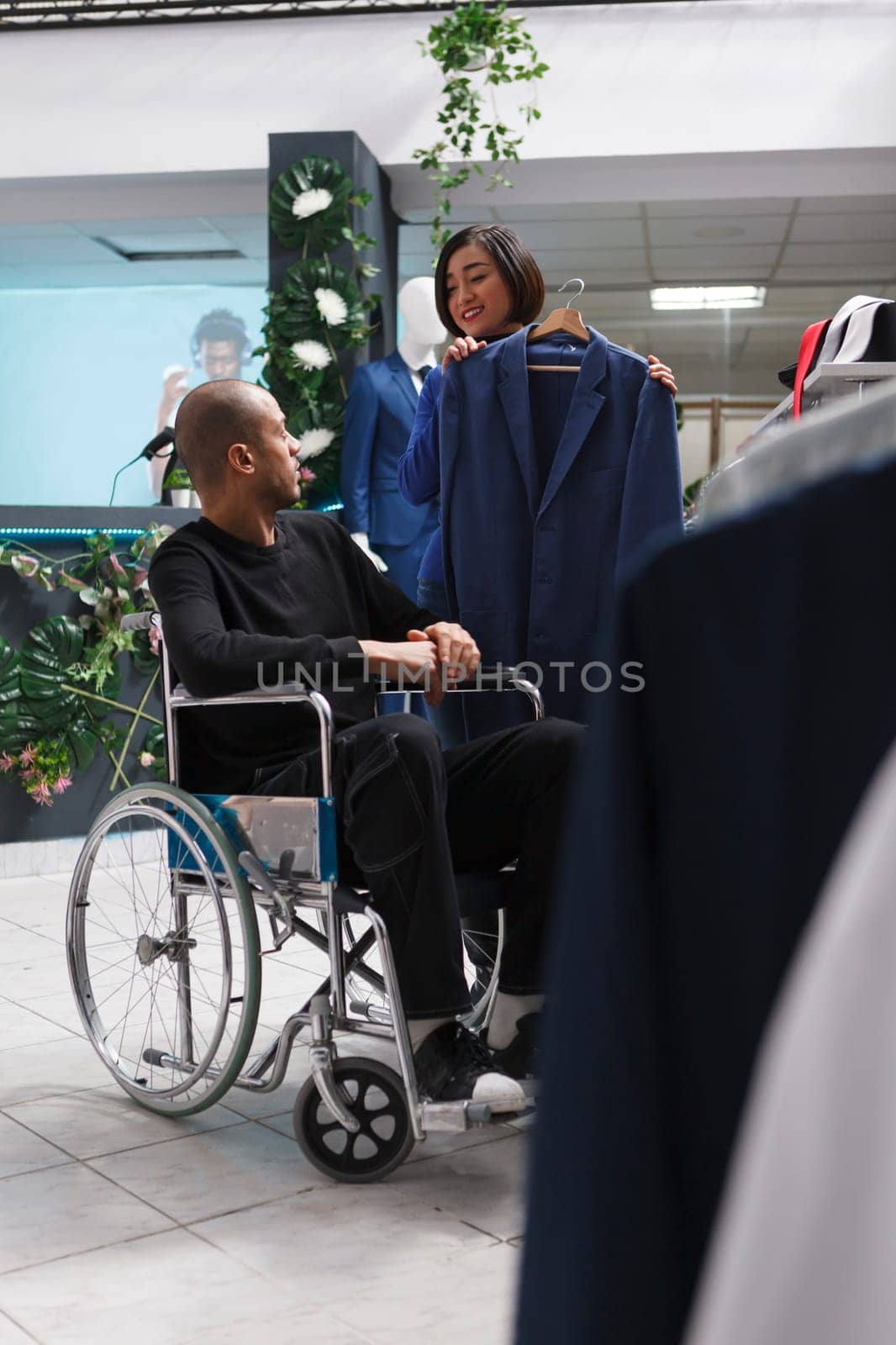 Clothing store asian woman employee providing support to customer with physical impairment in selecting jacket. Mall worker consulting arab man in wheelchair in choosing formal outfit