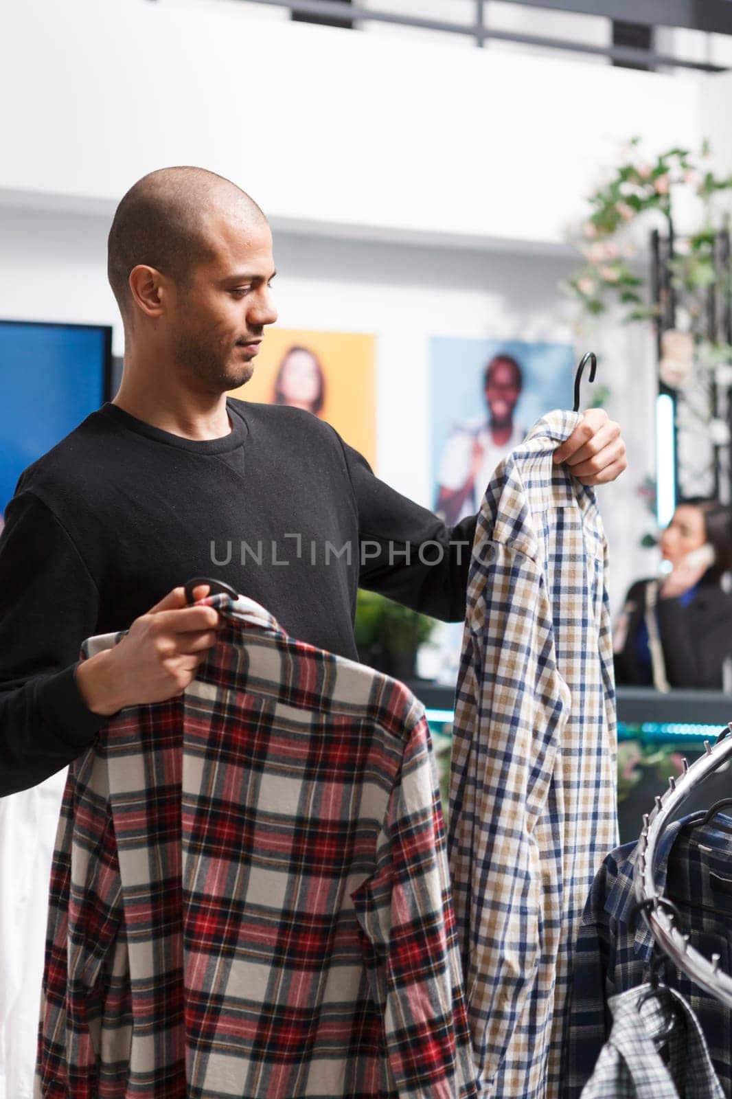 Shopping mall boutique arab man customer browsing plaid shirts and holding two outfits on hangers. Clothing store client choosing casual menswear apparel, considering style and fit
