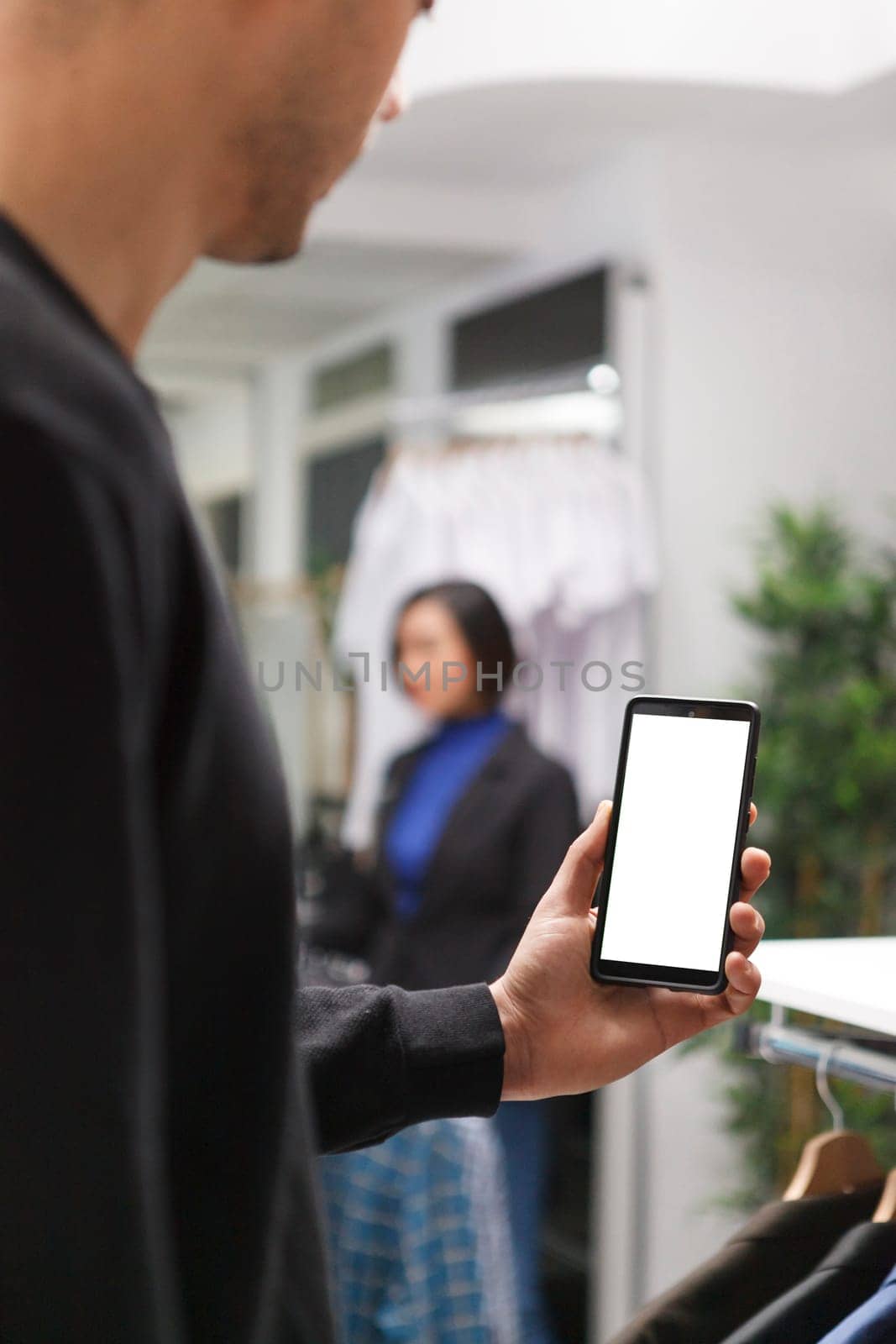Clothing store customer holding smartphone with blank screen mockup while exploring apparel online. Boutique man hand using mobile phone with empty white display while shopping in mall