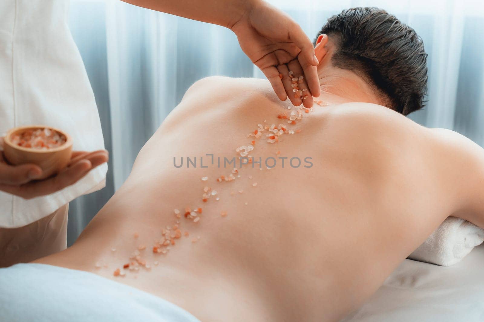 Blissful man customer having exfoliation treatment in luxury spa salon with warmth candle light ambient. Salt scrub beauty treatment in health spa body scrub. Quiescent