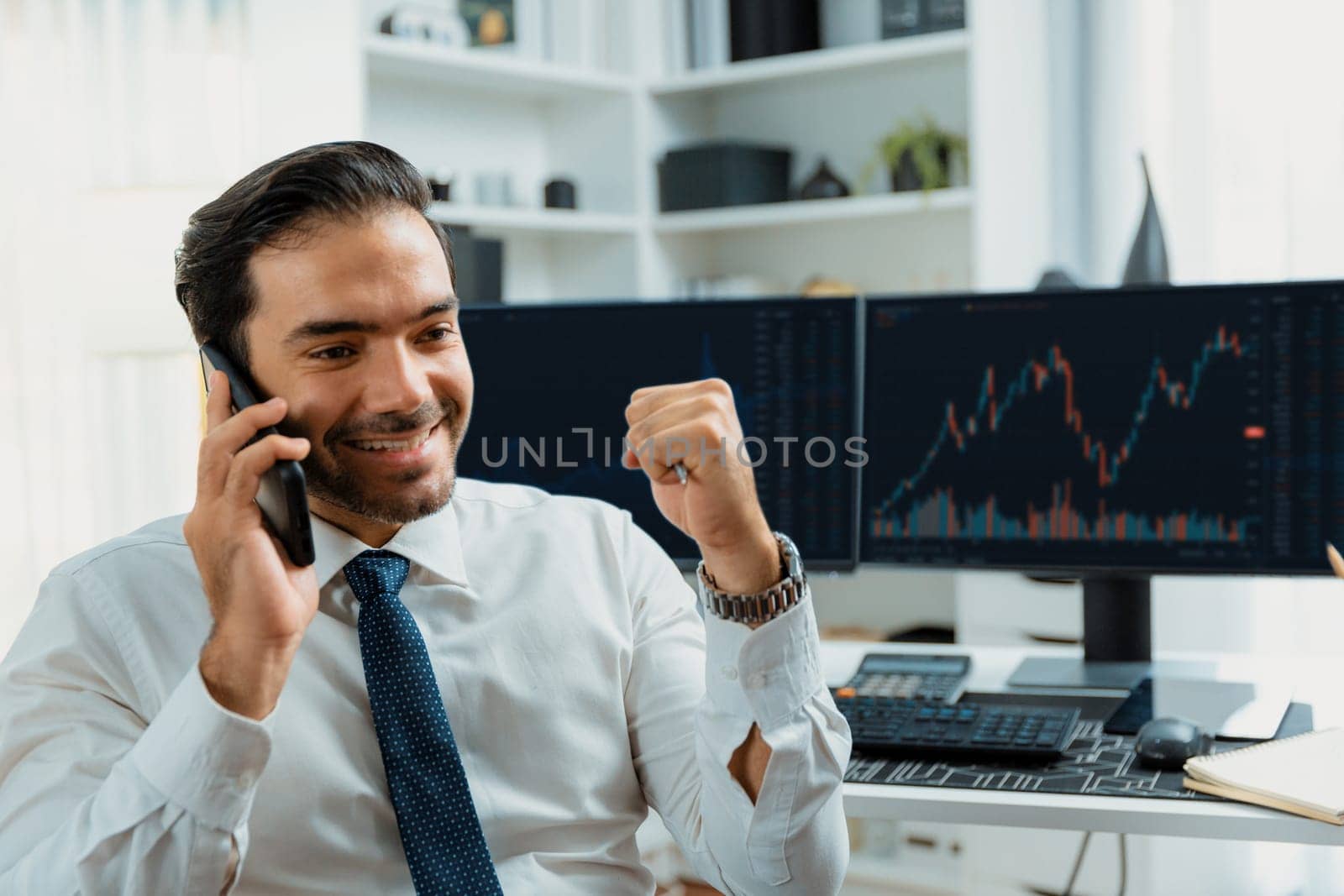 Successful in smiling trader businessman raising fist up, calling on phone against stock exchange investment analysis screen to invest high profit financial technology market at home office. Surmise.