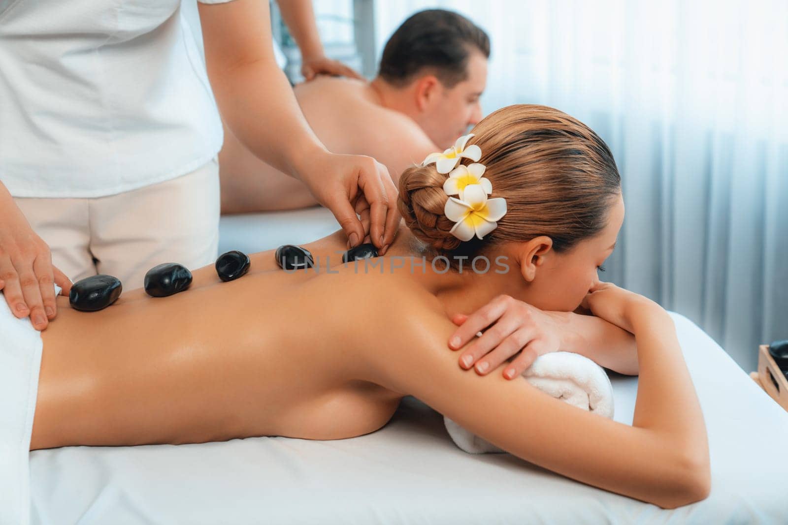 Hot stone massage at spa salon in luxury resort with day light serenity ambient, blissful couple customer enjoying spa basalt stone massage glide over body with soothing warmth. Quiescent