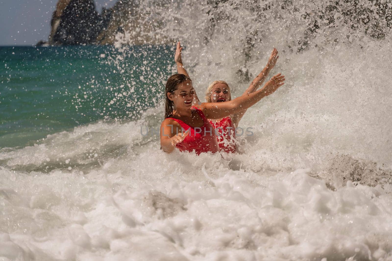 Women ocean play. Seaside, beach daytime, enjoying beach fun. Two women in red swimsuits enjoying themselves in the ocean waves and raising their hands up. by Matiunina