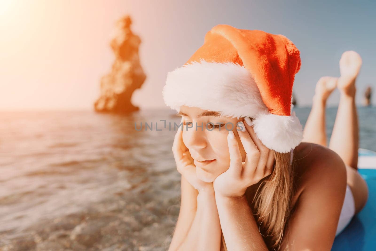 Woman sea sup. Close up portrait of happy young caucasian woman with long hair in Santa hat looking at camera and smiling. Cute woman portrait in a white bikini posing on sup board in the sea by panophotograph