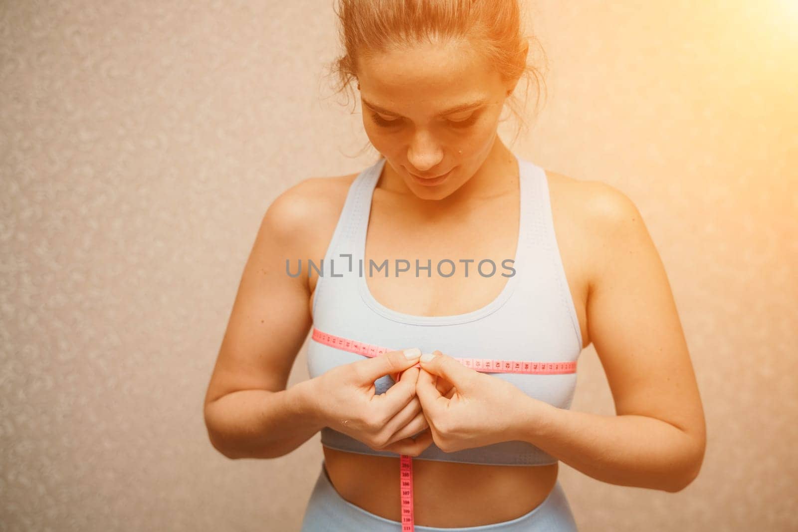 Cropped view of slim woman measuring breasts with tape measure at home, close up. A European woman checks the result of a weight loss diet or liposuction indoors. Healthy lifestyle. by Matiunina